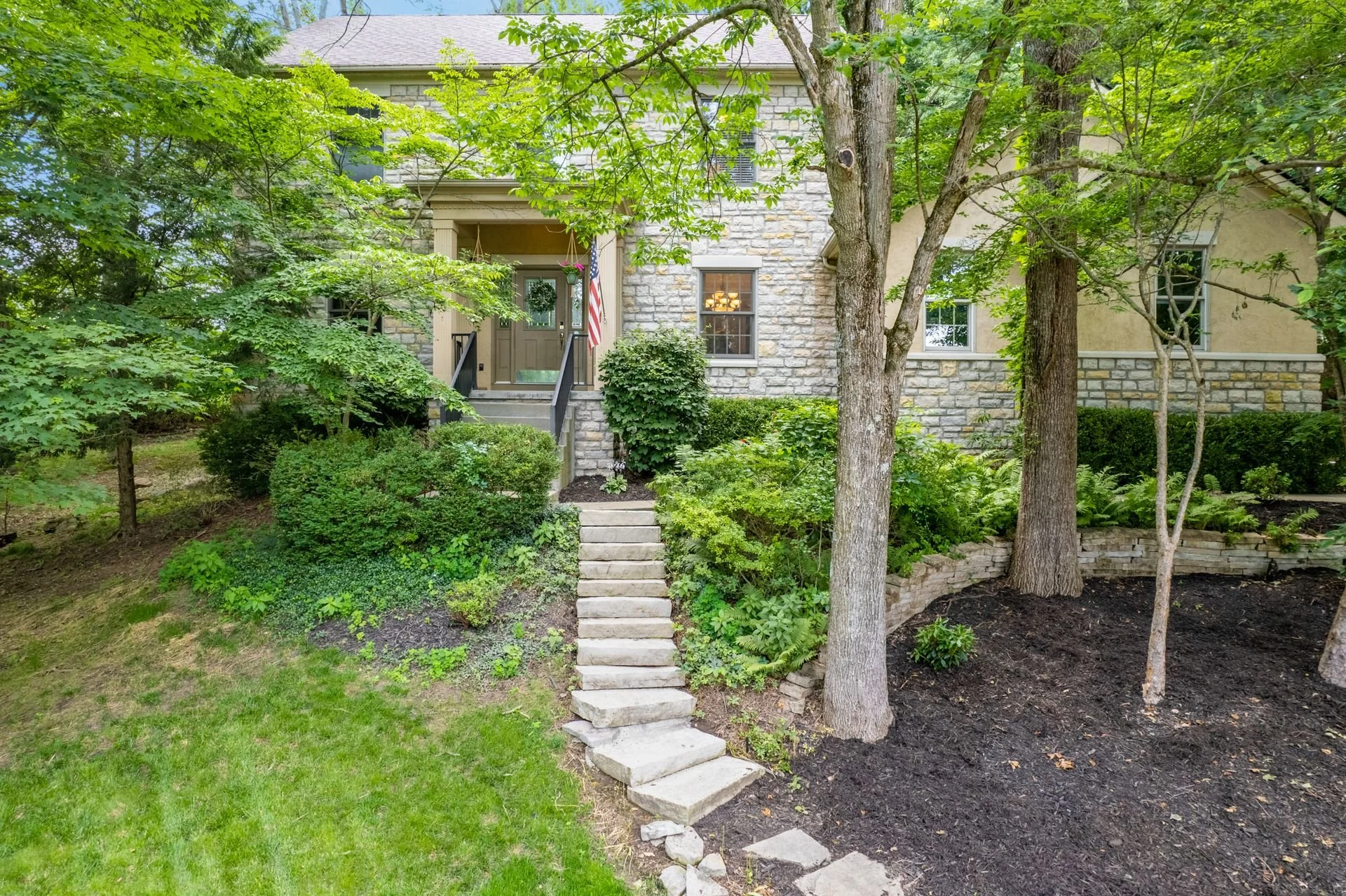 A house with a stone and stucco exterior, surrounded by lush green trees and shrubs, with a stone staircase leading to the front door, which is decorated with an American flag and hanging flower baskets.