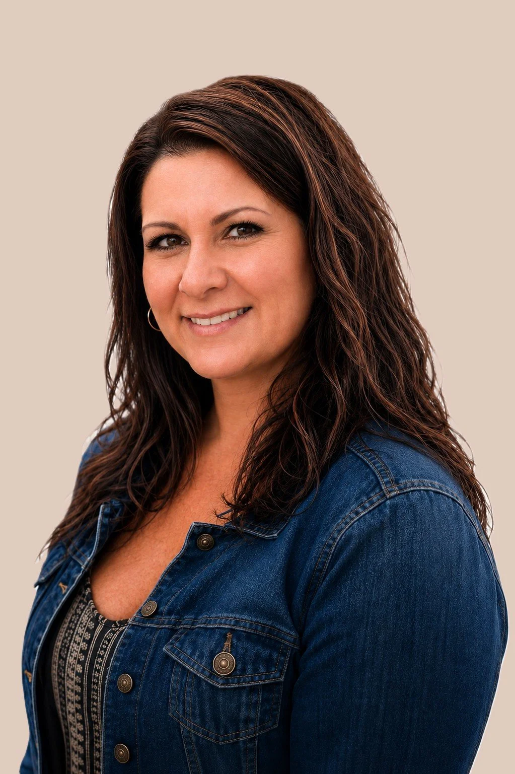 A woman with long, wavy dark brown hair smiling, wearing a denim jacket and a patterned top against a beige background.