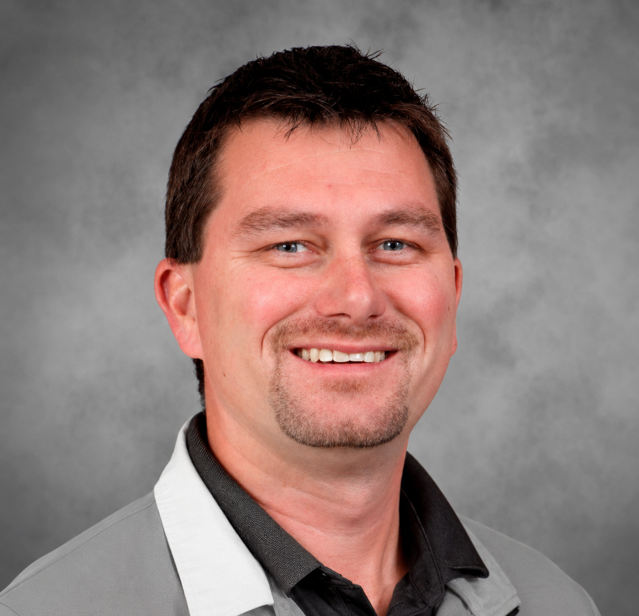 Portrait of a smiling man with short dark hair, blue eyes, and facial hair, wearing a gray collared shirt with a black undershirt, against a gray background.