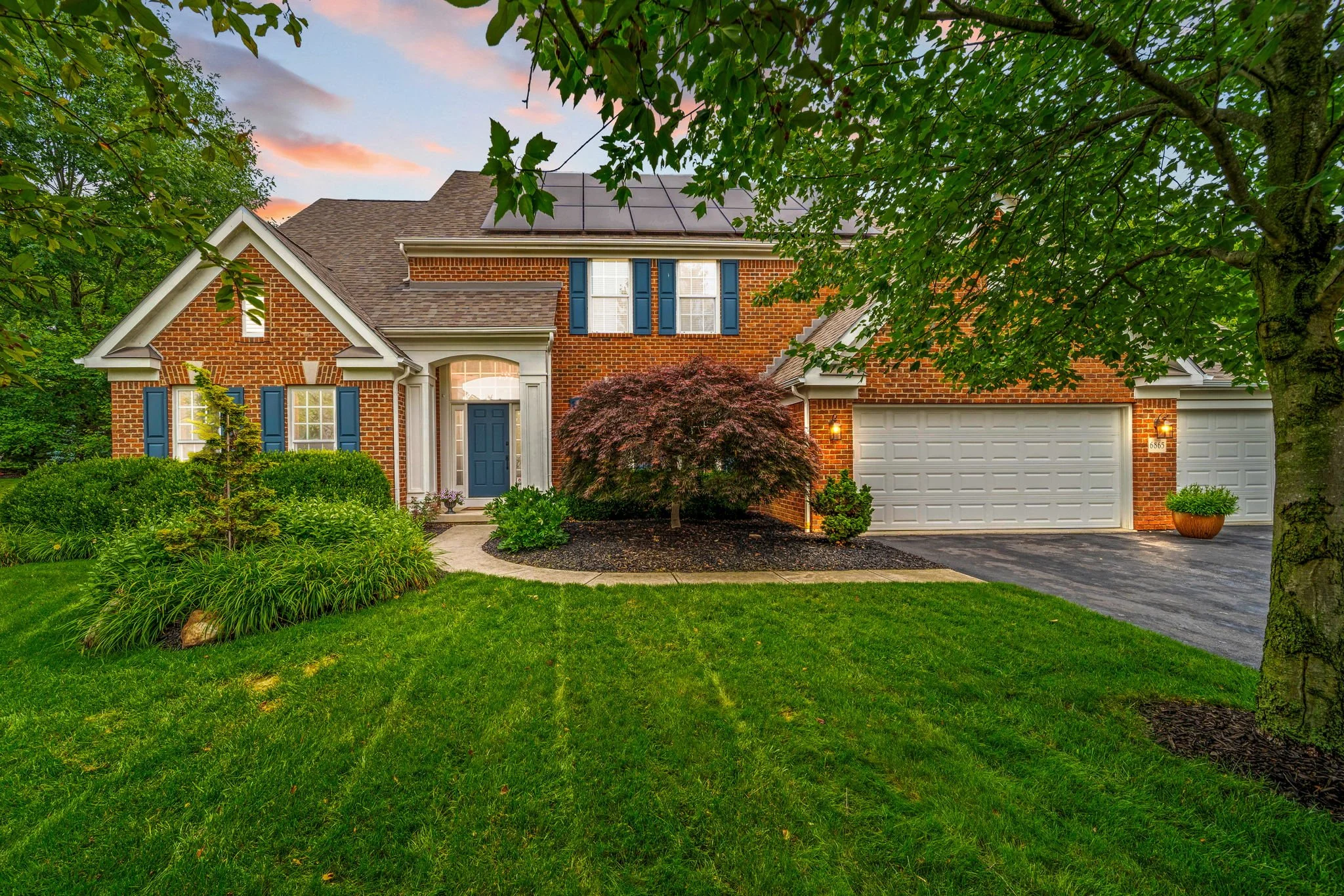 A two-story brick house with dark blue shutters and a dark blue front door, surrounded by green trees and shrubs, with a paved driveway and a white garage door, set against a colorful sunset sky.