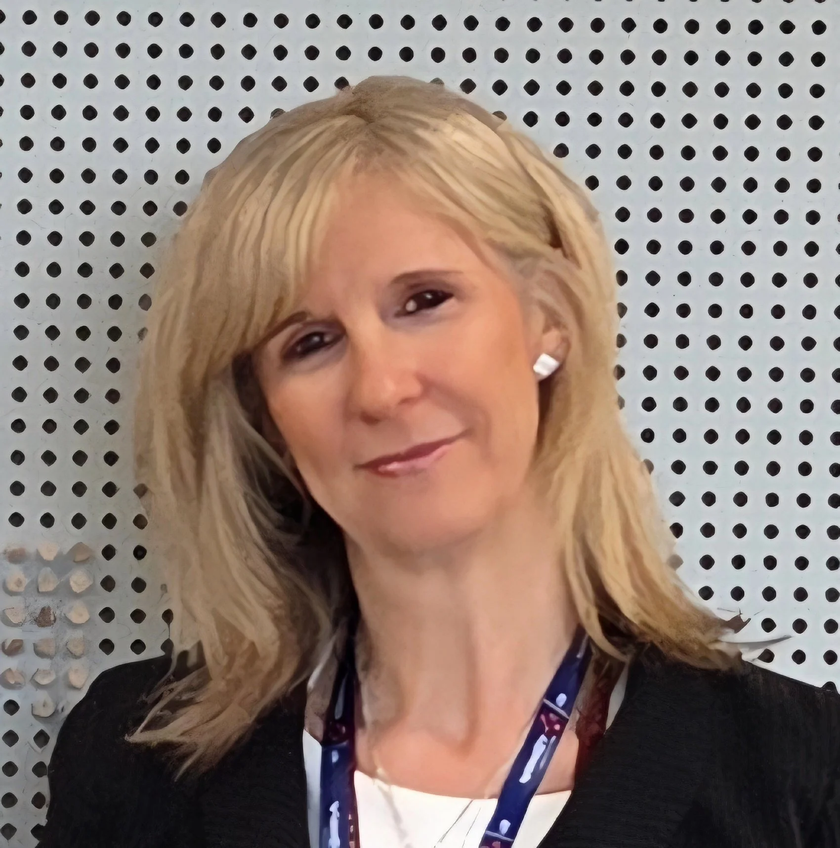 Close-up photo of a woman with long blonde hair, wearing earrings and a dark top, standing in front of a white pegboard with black holes.