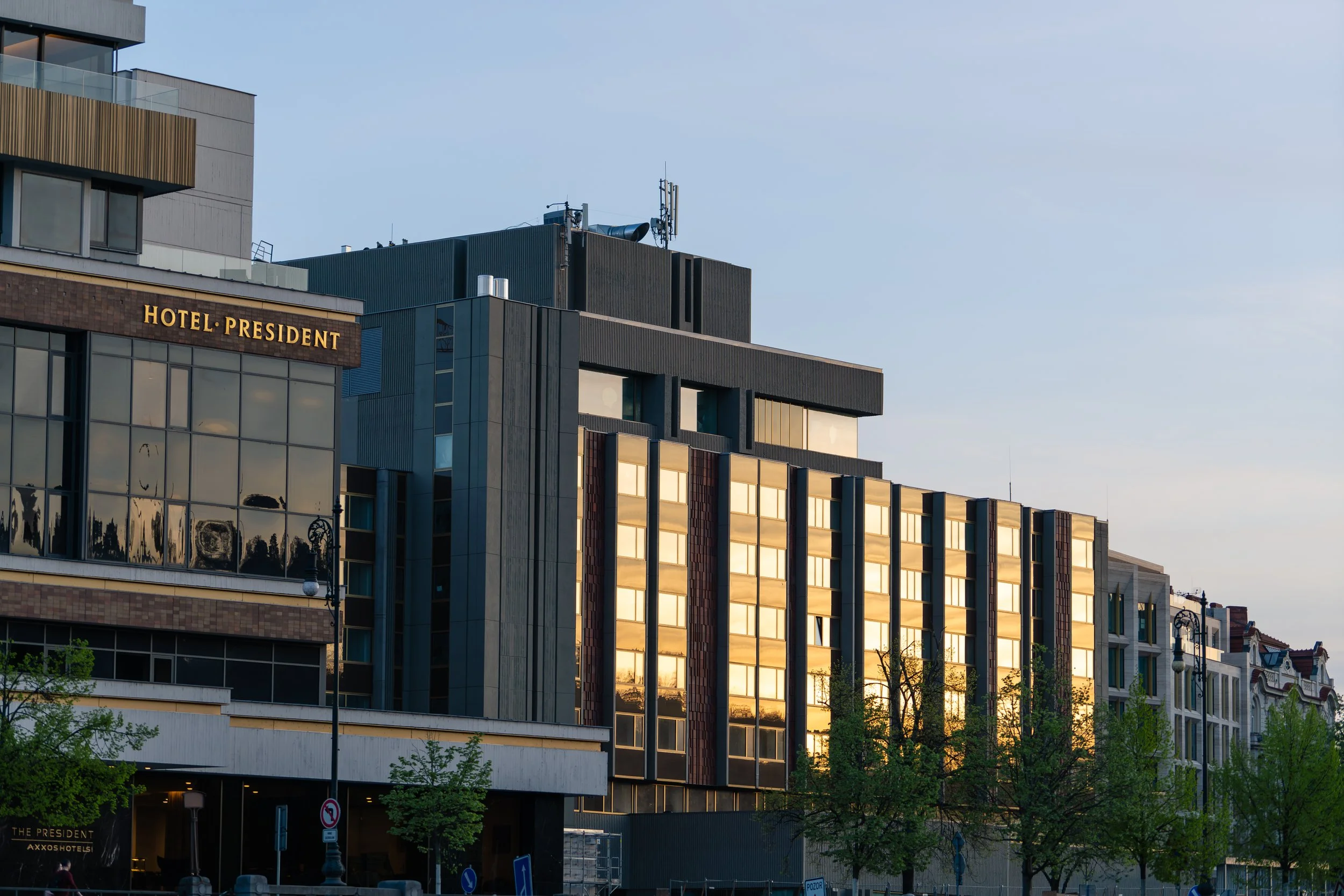 Mehrstöckiges modernes Hotelgebäude namens 'Hotel President' mit reflektierenden Glasfenstern und einem Restaurantbereich im Erdgeschoss, umgeben von Straßen und Bäumen bei Sonnenuntergang.