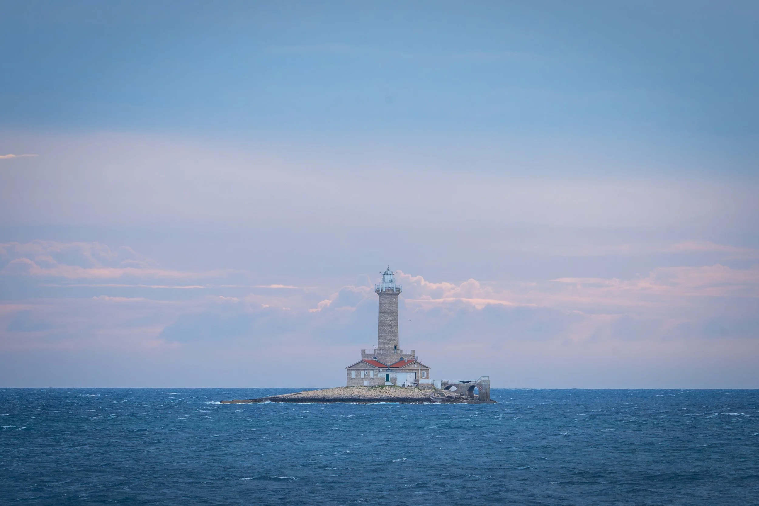Leuchtturm auf einer kleinen Insel im Meer, bei bewölktem Himmel.