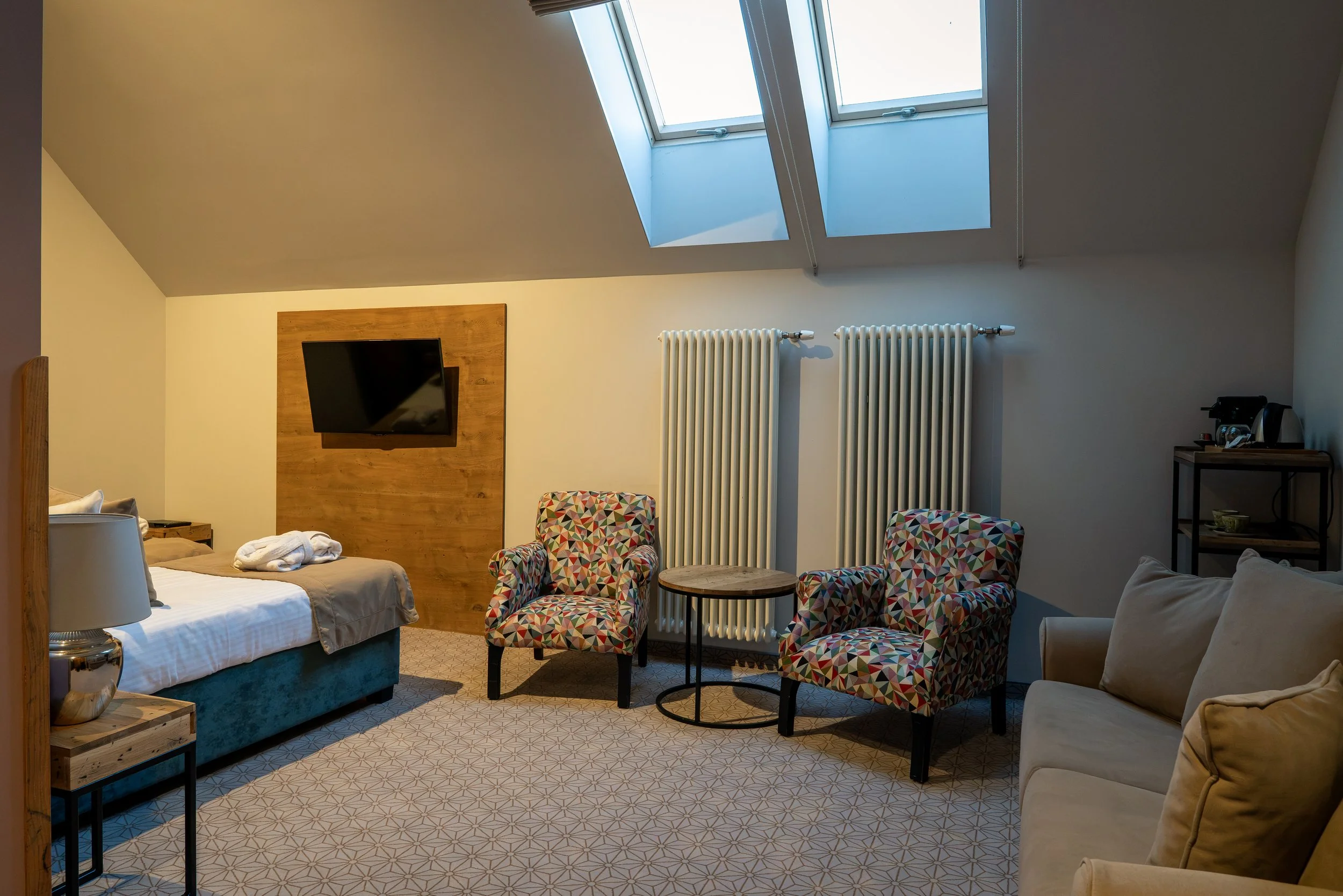 Ein Hotelzimmer mit Dachfenstern, einem Bett, einem Fernseher, einem Stuhl mit buntem Muster, einer Heizung, einem kleinen Tisch, einem Nachttisch mit einer Lampe und einem kleinen Kaffeekocher.