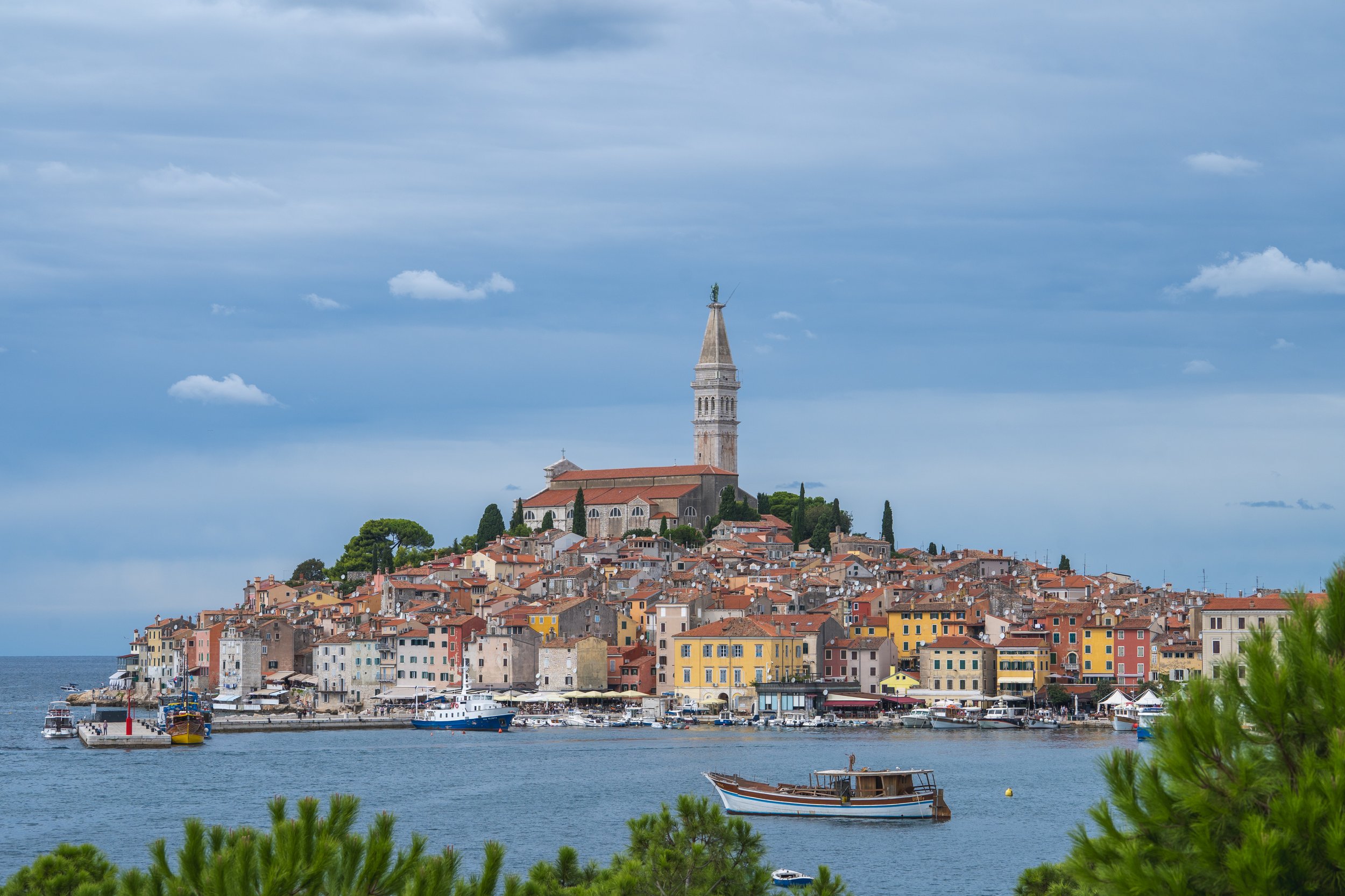 Eine Küstenstadt mit bunten Häusern, ein Schloss auf einem Hügel mit einer Kirche, und Boote im Wasser bei bewölktem Himmel.