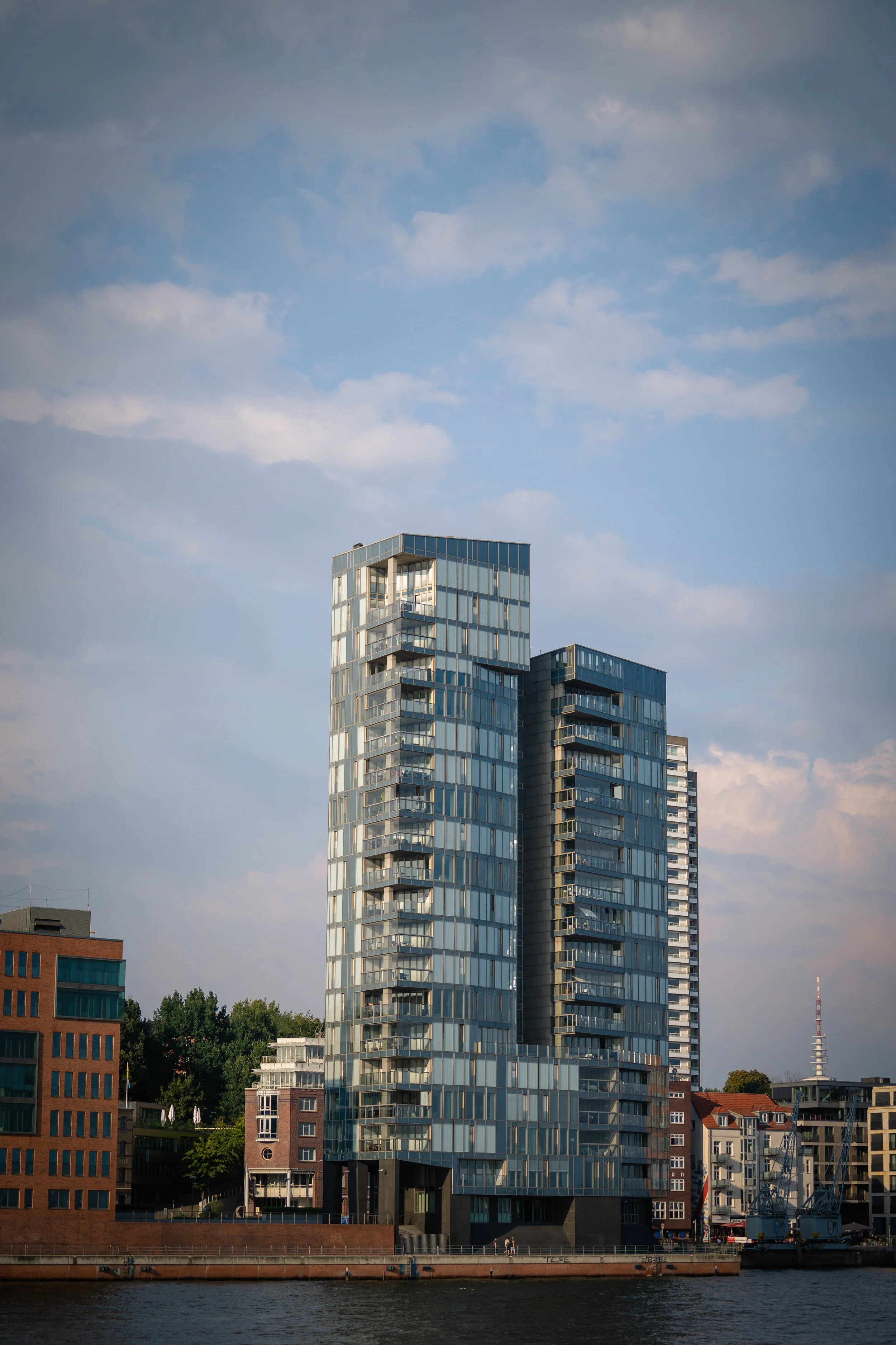 Moderne hohe Bürogebäude mit vielen Fenstern am Fluss, im Hintergrund ein Fernsehturm und ein blauer Himmel mit Wolken.