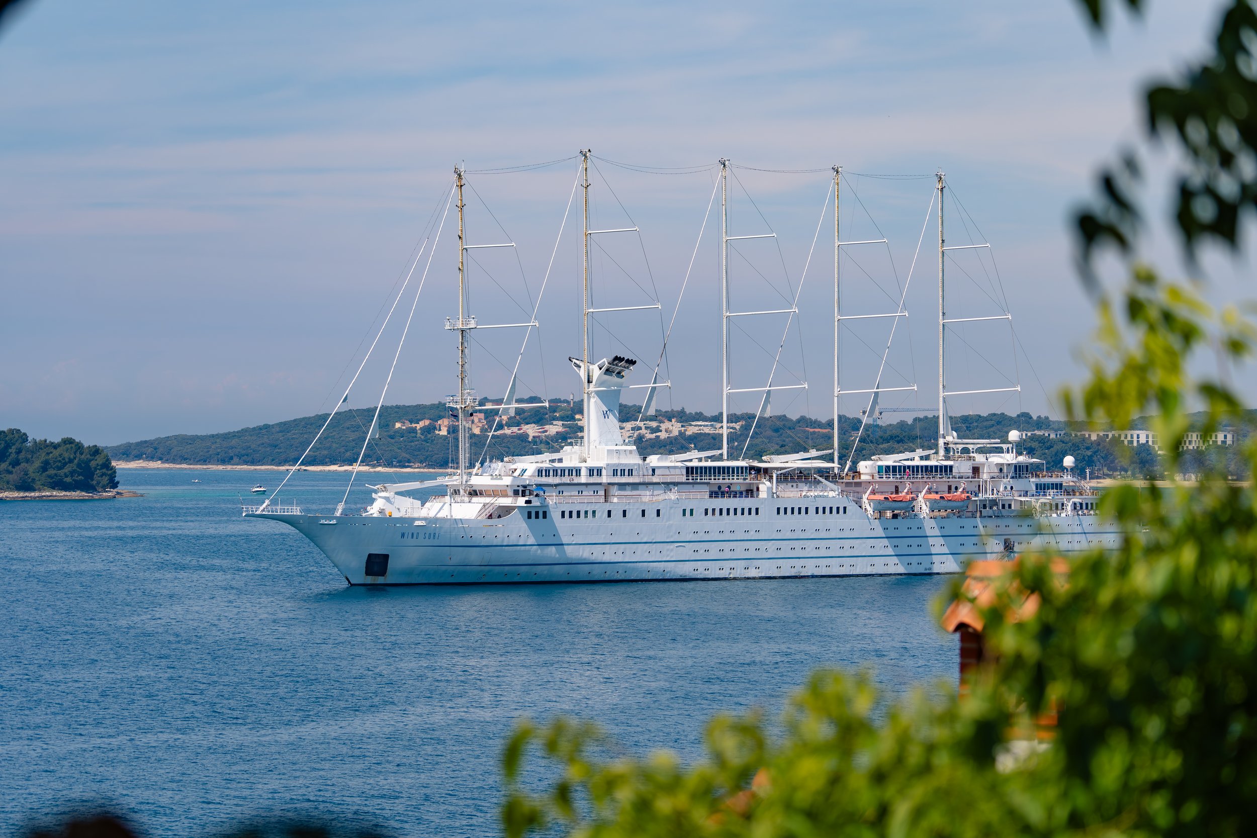 Ein weißes Kreuzfahrtschiff namens Wind Surf liegt auf dem Meer, im Hintergrund sind Hügel und Häuser sichtbar. Blätter im Vordergrund sind unscharf.