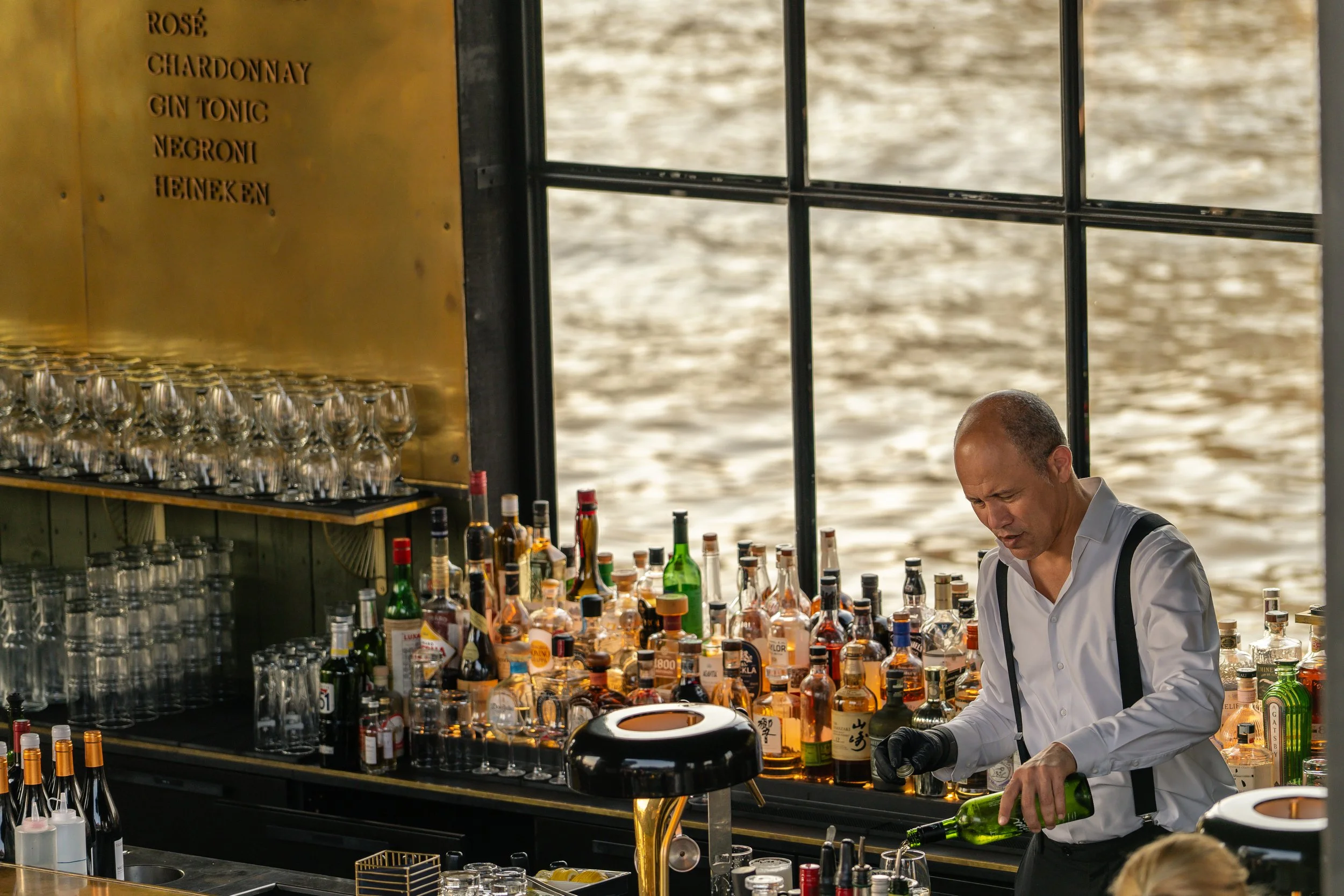 Barkeeper bereitet eine Bar an einem Wasserweg vor, mit vielen Flaschen und Gläsern, während im Hintergrund die Wasseroberfläche sichtbar ist.