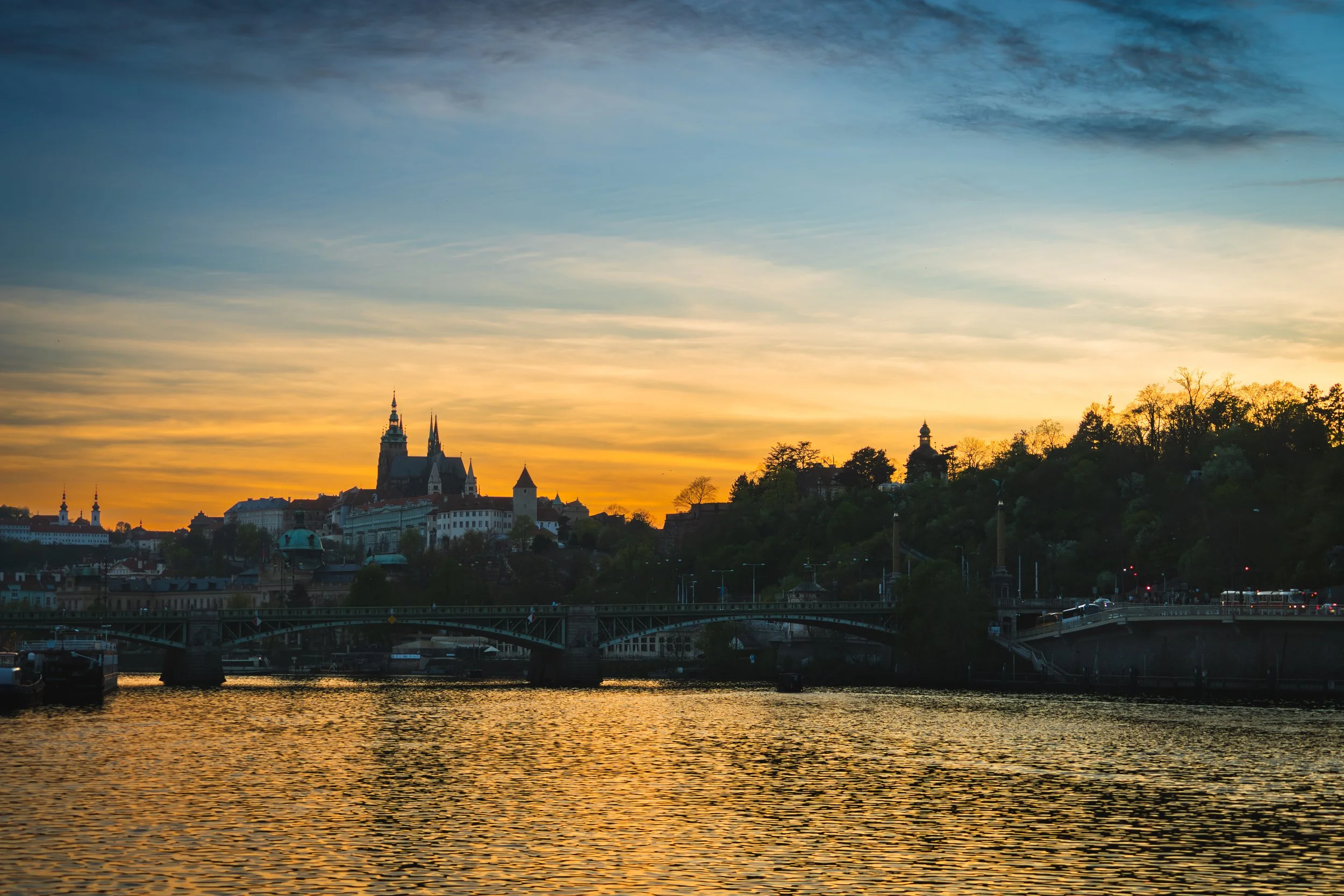 Blick auf eine Stadt bei Sonnenuntergang mit einer Brücke, Wasser im Vordergrund und historischen Gebäuden mit Türmen und Kirchen im Hintergrund.