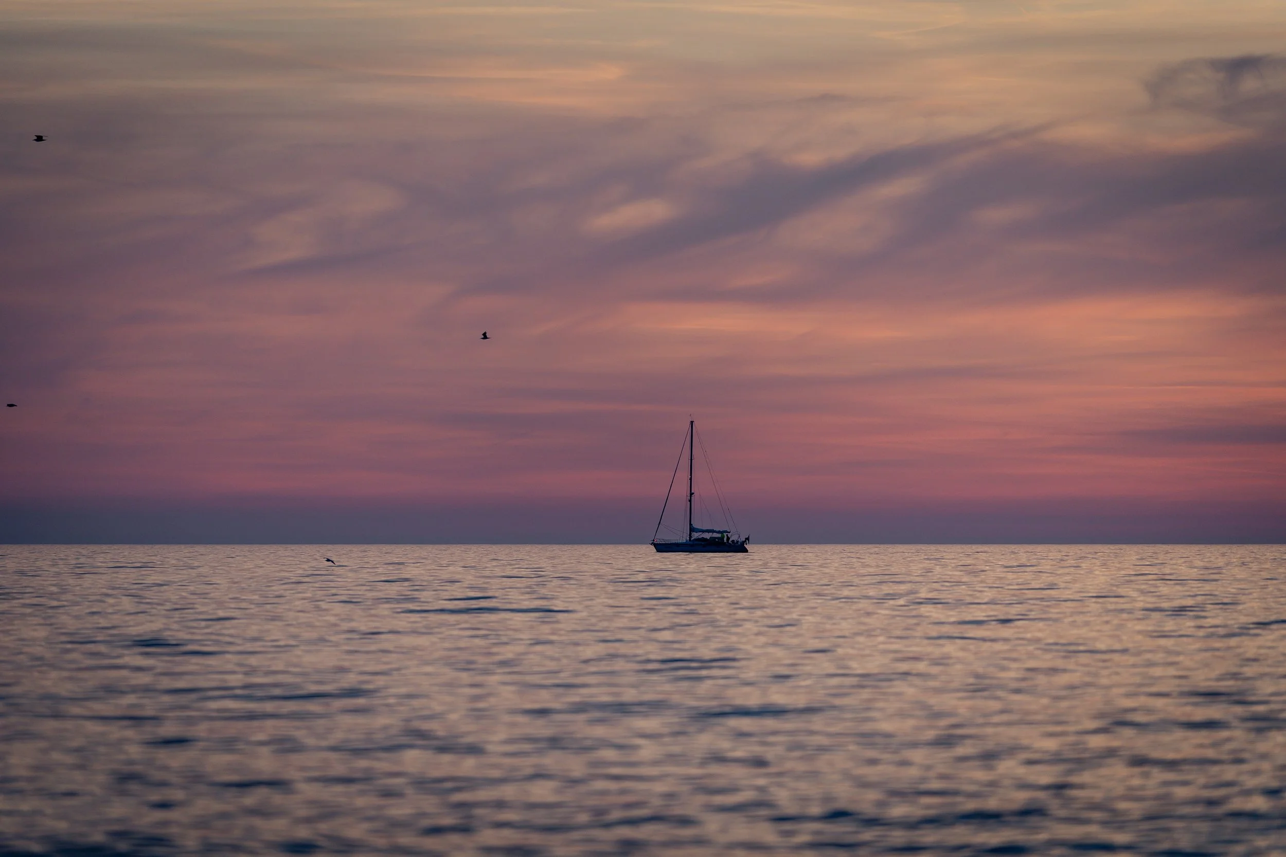 Segelboot auf ruhigem Meer bei Sonnenuntergang mit farbigem Himmel und fliegenden Vögeln.