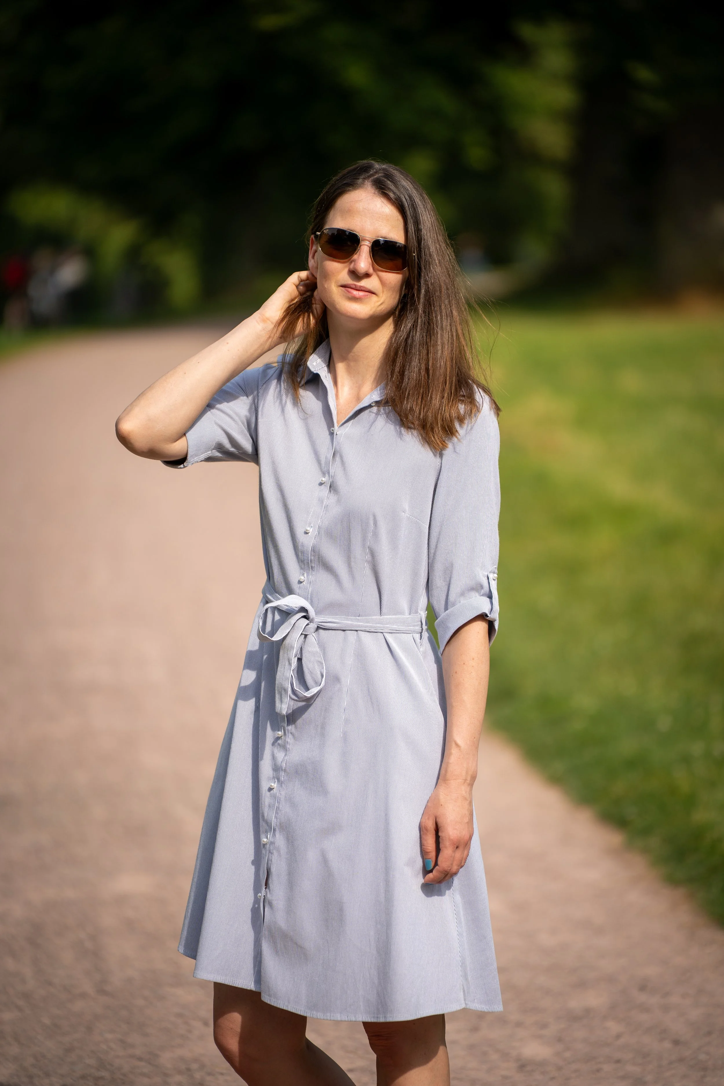 Frau in einem gestreiften, hellblauen Kleid mit Hemdkragen und Knöpfen steht auf einem Weg im Grünen, trägt Sonnenbrille und hat braune, schulterlange Haare.