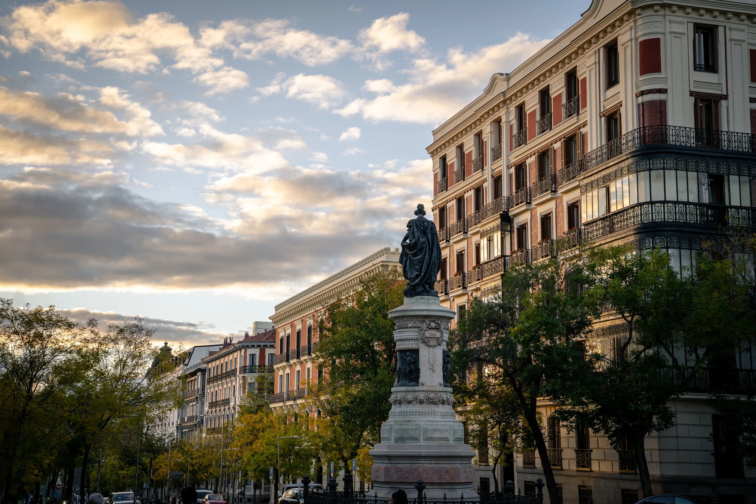Historische Straße mit mehrstöckigen Gebäuden, Statue auf einem Sockel im Vordergrund, Baumkronen und Wolken im Himmel bei Sonnenuntergang.