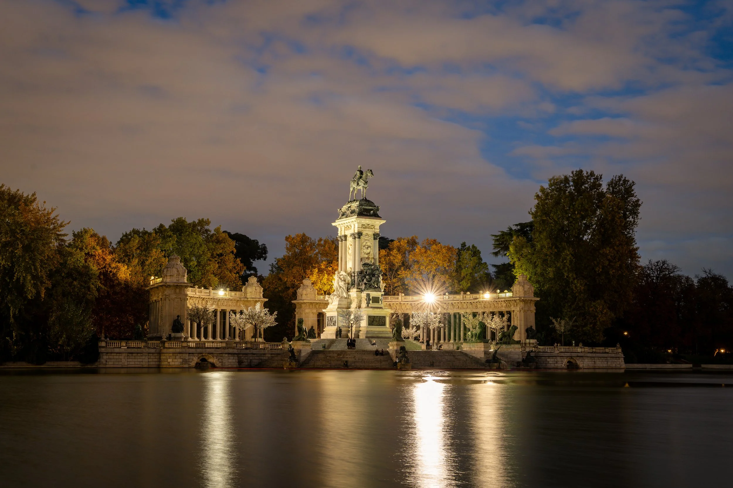 Der Eiserne Kaiser auf einem hohen Sockel in einem Park mit vielen Bäumen. Der Himmel ist bewölkt, und das Wasser im Vordergrund spiegelt die Szene wider.