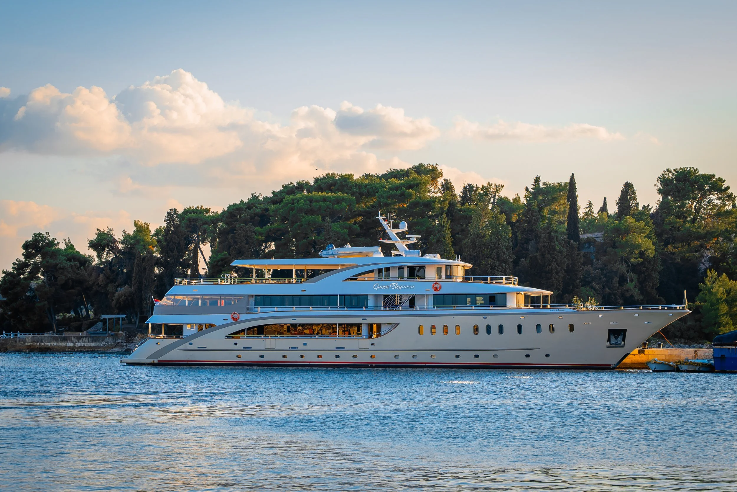 Ein großer weißer Yacht namens 'Queen Eleganza' liegt im Wasser neben einer Uferpromenade, vor einem Hintergrund voller grüner Bäume und einem Himmel mit weißen Wolken.
