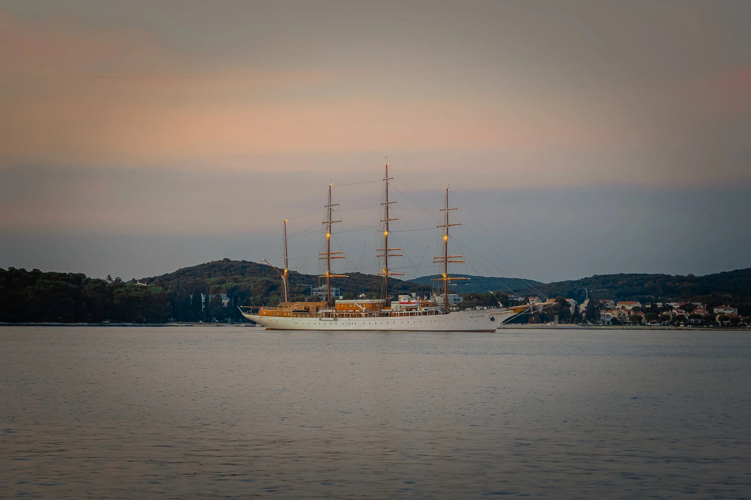 Ein großer weißer Segelschiff mit drei Masten segelt auf ruhigem Wasser bei Sonnenuntergang, mit Stadt und Hügeln im Hintergrund.
