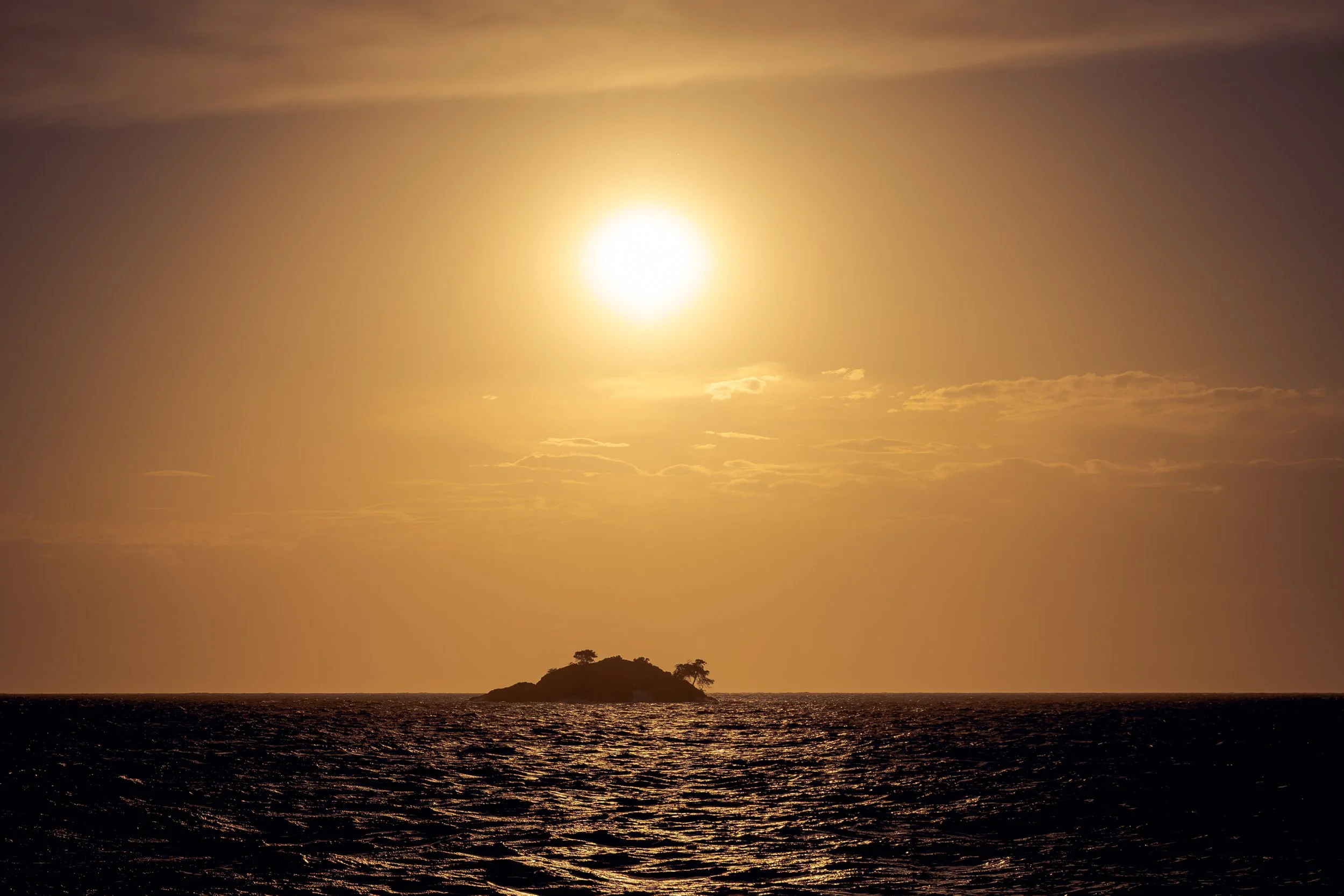 Sonnenuntergang über dem Meer mit einer kleinen Insel im Vordergrund, der Himmel ist gelblich-orange mit wenigen Wolken.