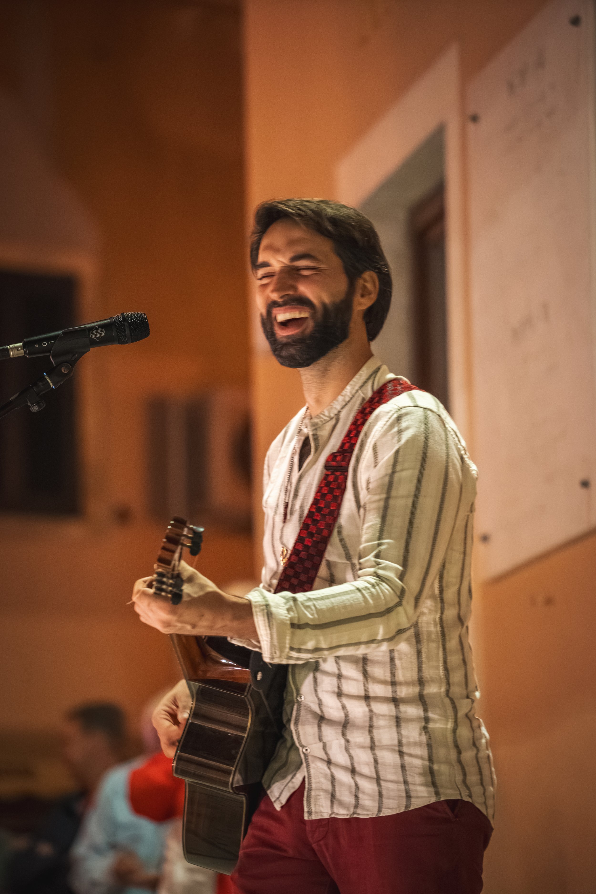 Ein lachender Mann mit dunkler Haut, Bart und dunklem Haar, spielt Gitarre und singt in einem Raum mit warmem Licht, im Hintergrund eine Person mit blauer Kleidung.