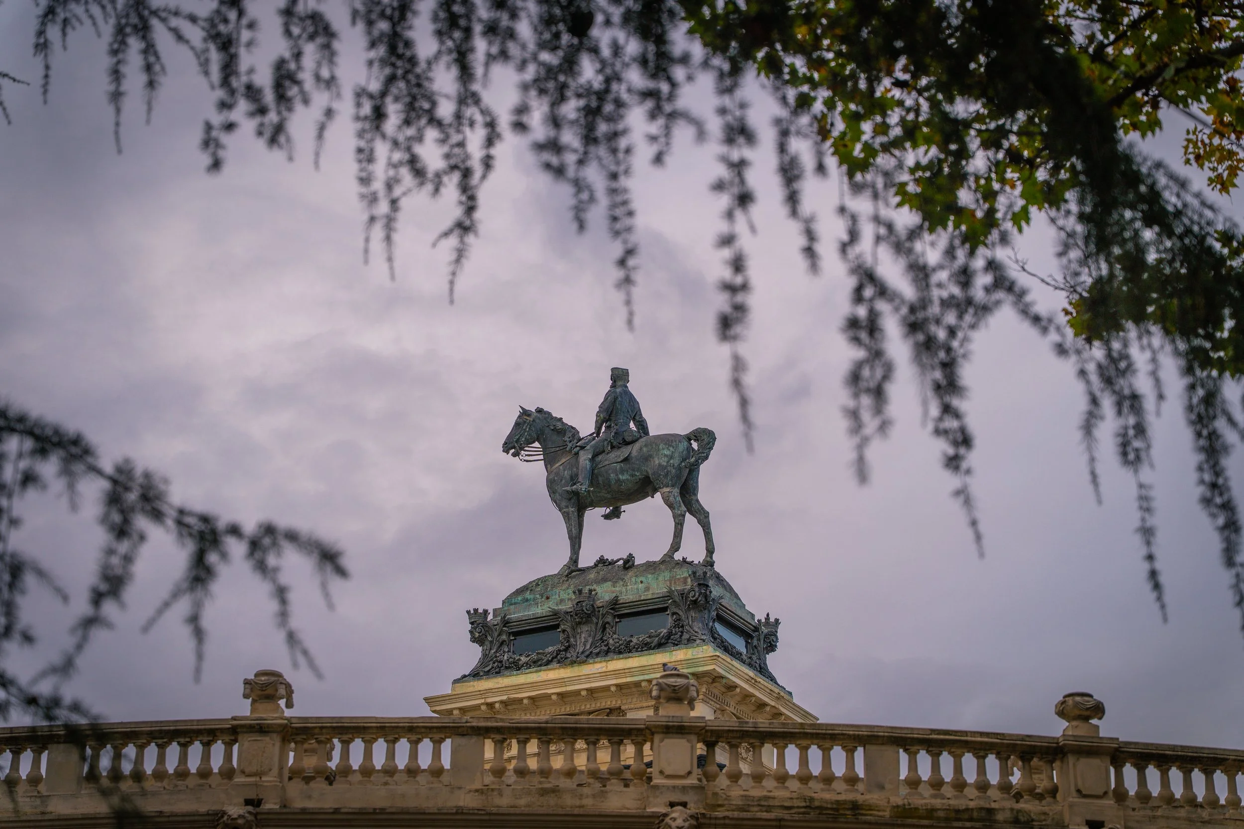 Den Blick nach oben auf eine barocke Brunnenskulptur mit Reiter und Pferd, umgeben von einer Balustrade, mit einem Himmel im Hintergrund, durch dessen Zwischenräume Zweige und Blätter eines Baumes sichtbar sind.