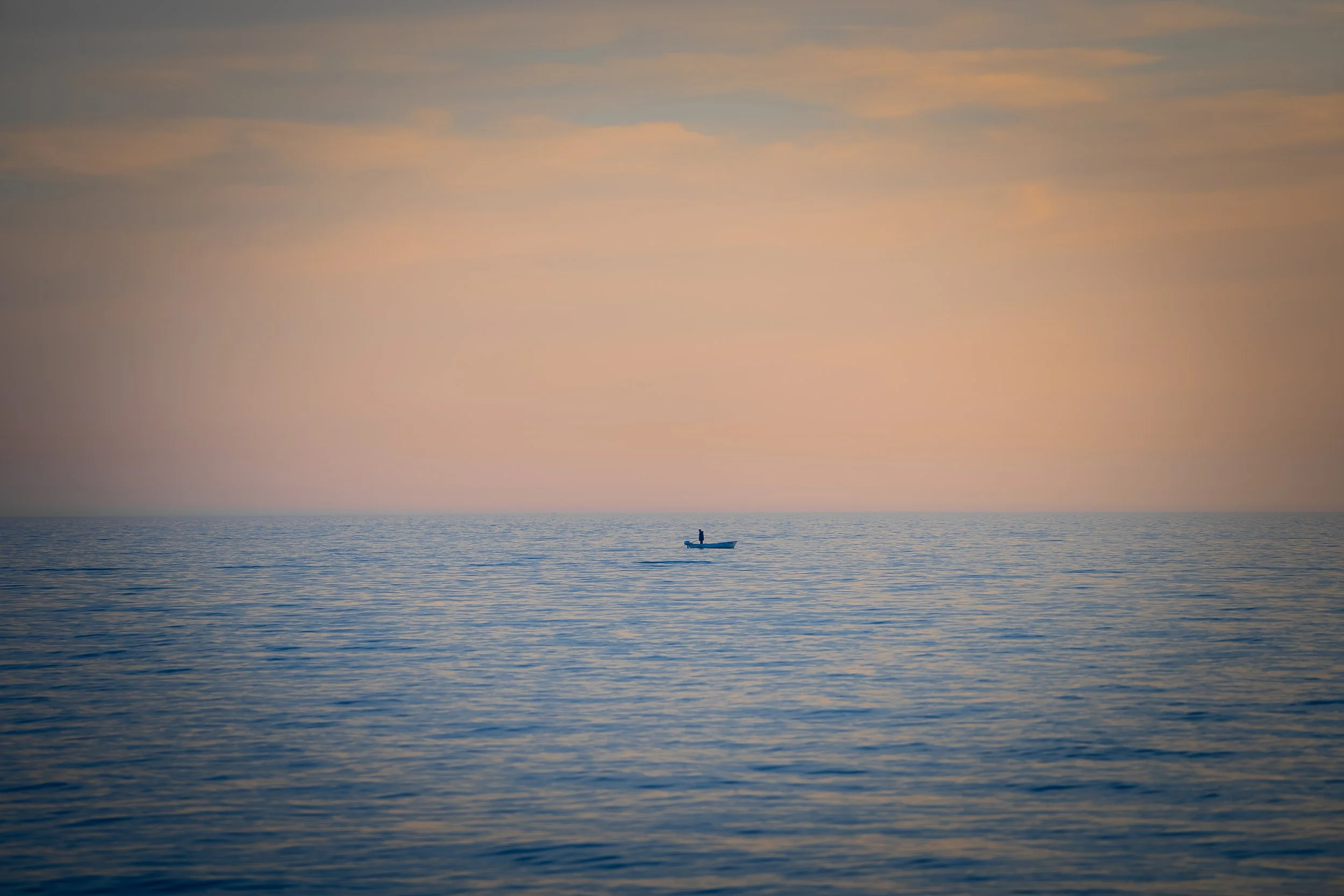 Ein einzelnes Boot mit einer Person an Bord auf ruhigem Meer während des Sonnenuntergangs, Himmel mit sanften Wolken in warmen Farbtönen.