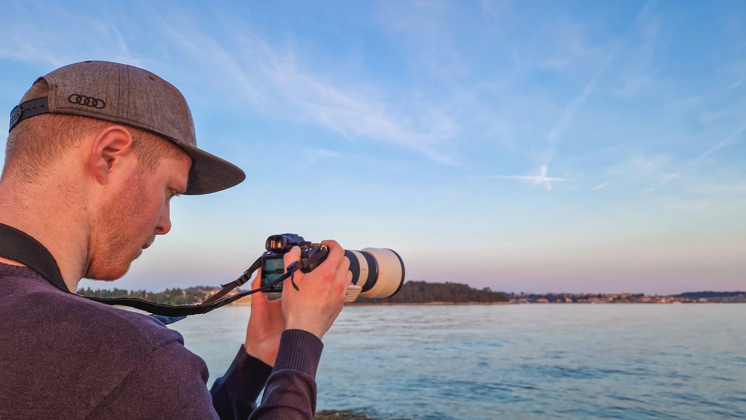 Ein Mann trägt eine graue Kappe mit einem schwarzen Logo, hält eine Kamera mit einem langen Teleobjektiv und fotografiert am Wasser bei Sonnenuntergang.