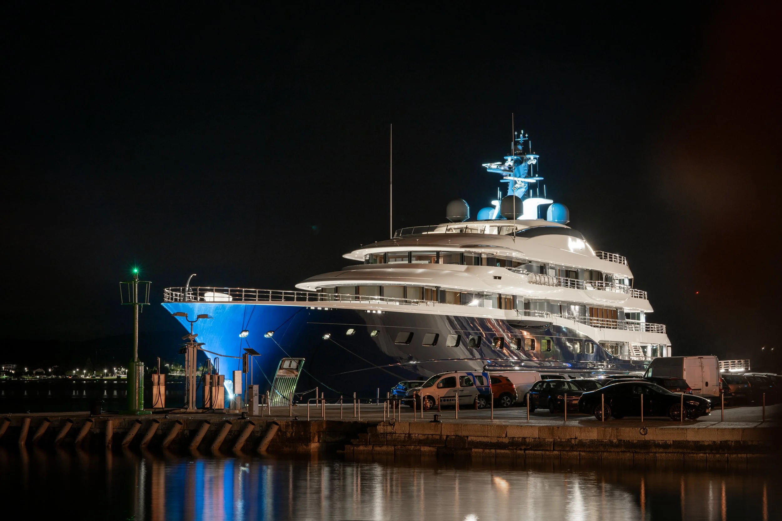 Ein großer, beleuchteter Yacht liegt nachts im Hafen, reflektiert im Wasser. Im Vordergrund sind das Wasser und einige Bojen sichtbar, der Himmel ist dunkel.