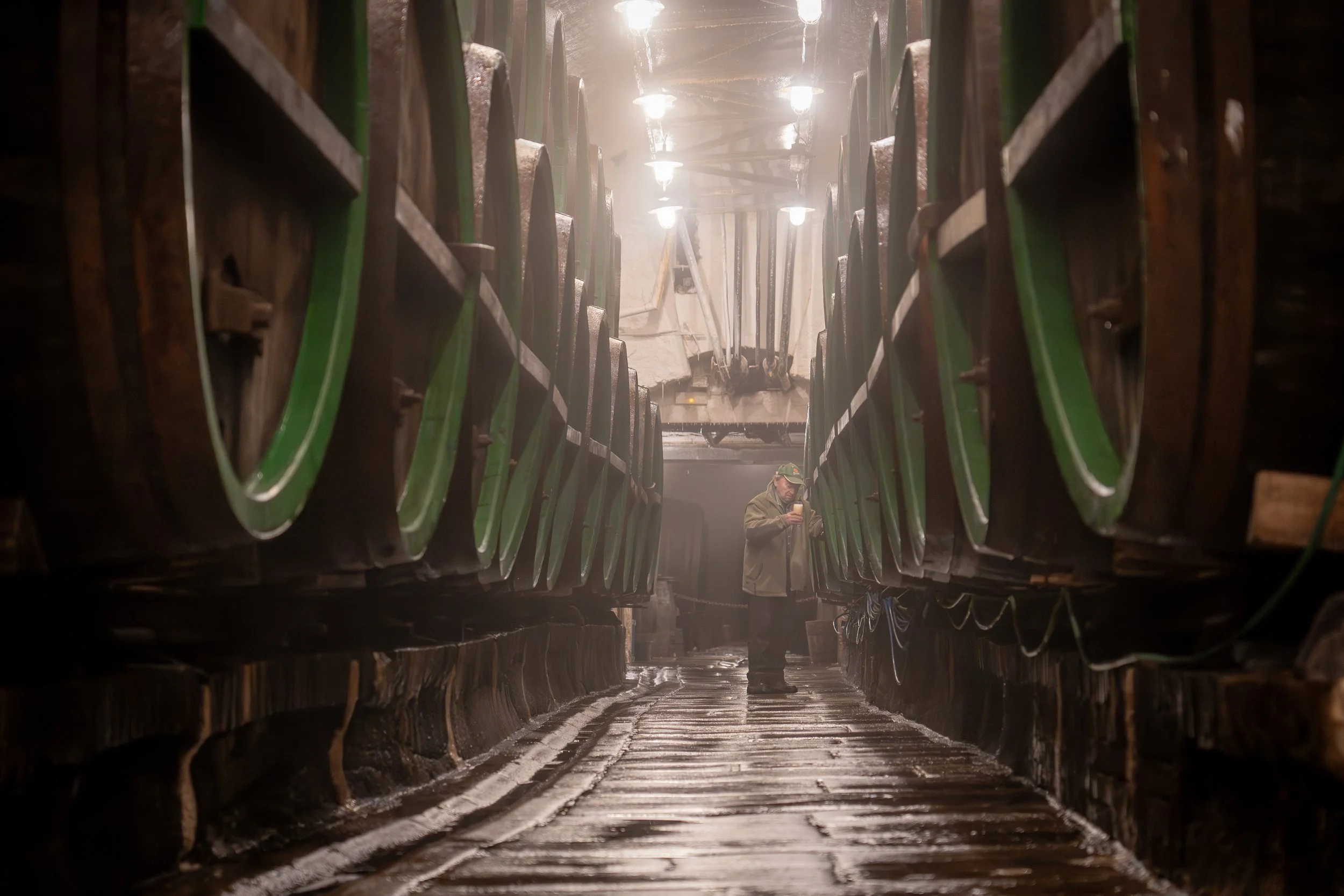 Ein Blick zwischen große Wagons in einer dunklen, industriellen Lagerhalle. Zwei Menschen stehen in der Mitte, eine Person hält ein Smartphone in der Hand. Das Licht hängt an der Decke, erzeugt eine diffuse Beleuchtung.