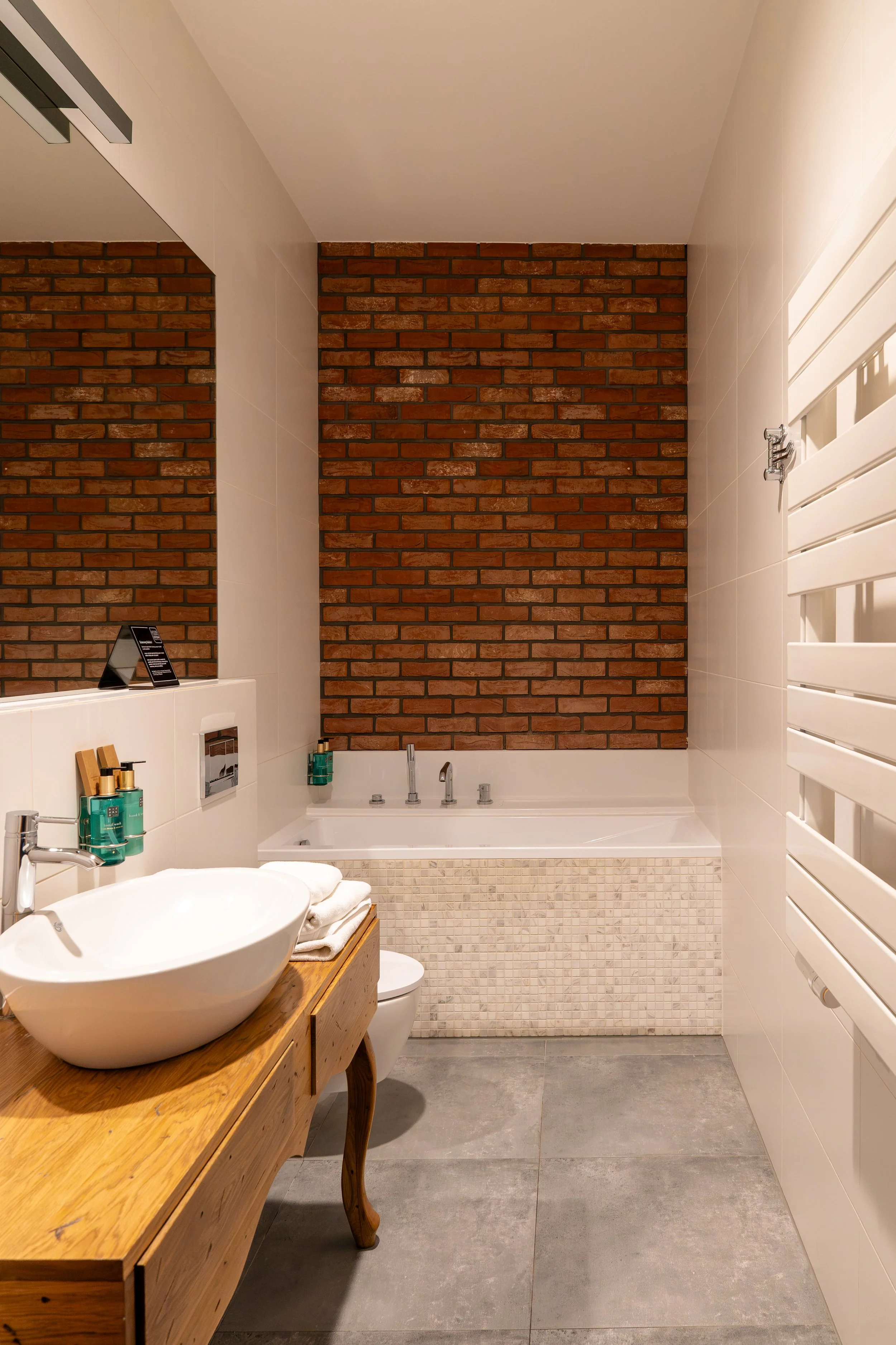 Ein modernes Badezimmer mit einer Badewanne, einem Waschbecken auf einem Holztisch, weißen Fliesen, einer roten Ziegelwand im Hintergrund und einem weißen Heizkörper an der rechten Wand.