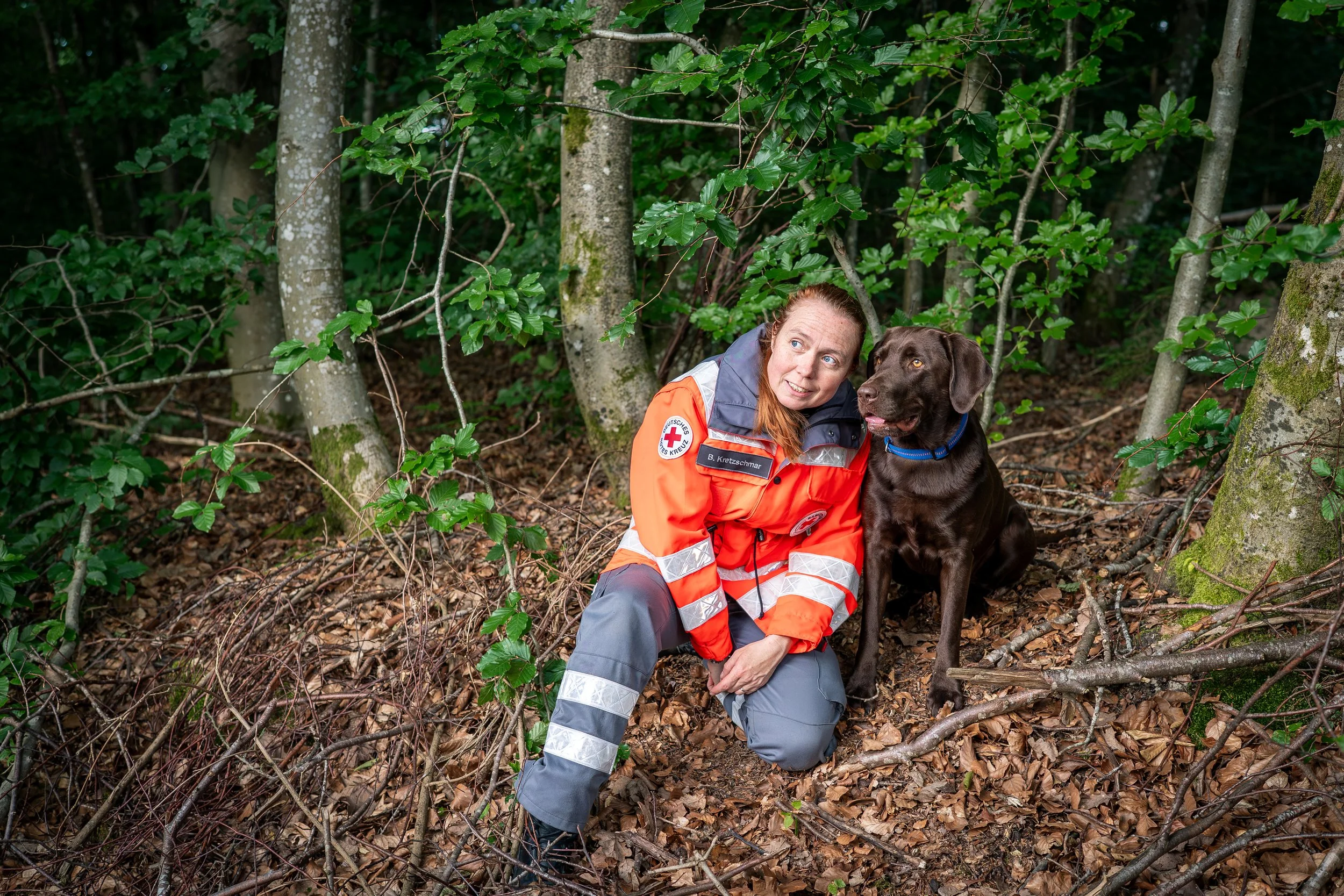 Eine rote Kreuz-Verbandsfleckenkraft in einem orangefarbenen Einsatzanzug mit Hund in einem Waldumfeld, umgeben von Bäumen und Laub