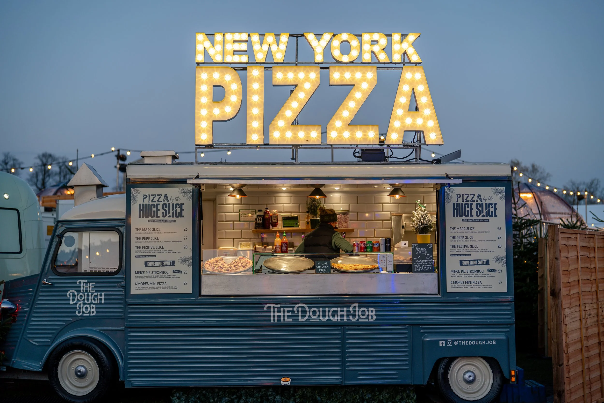 Ein Food-Truck mit großen Leuchtbuchstaben, die 'NEW YORK PIZZA' anzeigen, bei Dämmerung, mit Menütafeln und einem Mitarbeiter im Inneren.
