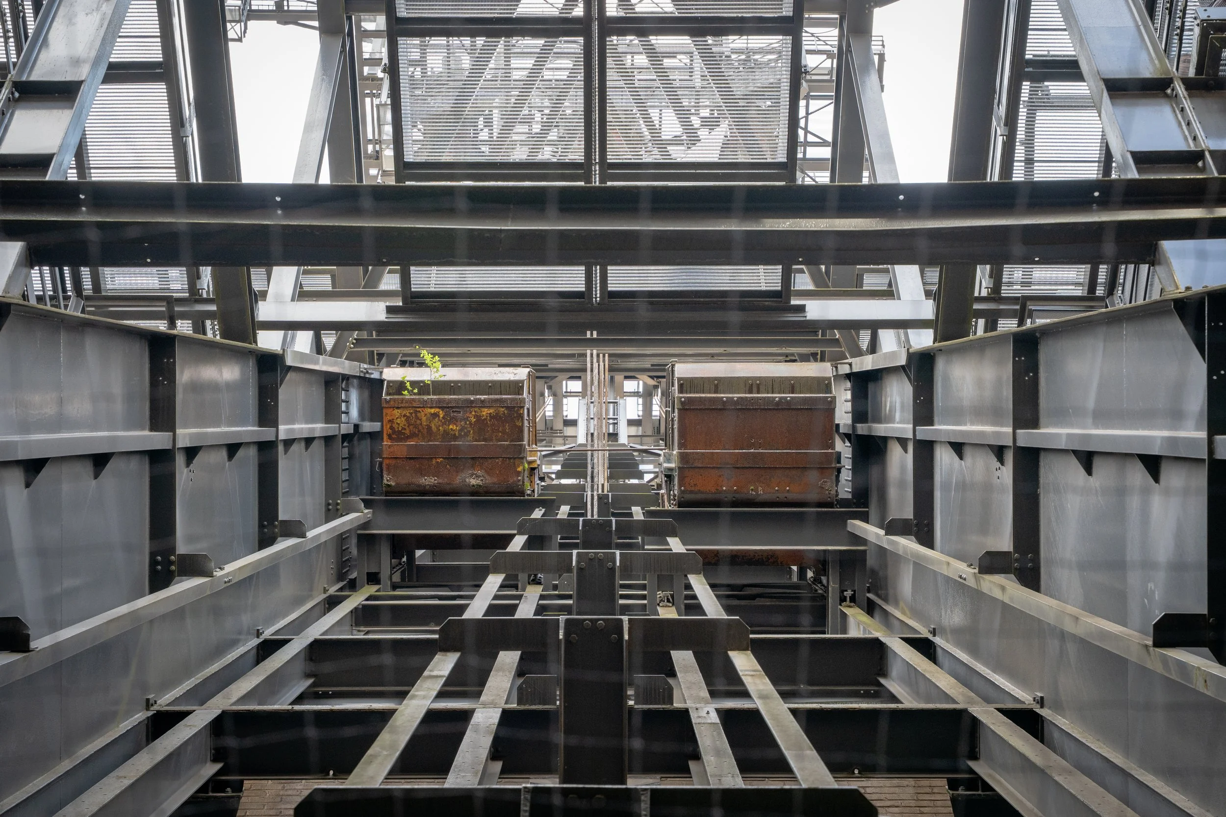 Blick nach oben auf eine moderne, industrielle Stahlkonstruktion mit Treppen, Gitterrosten und Rohren bei Tageslicht.