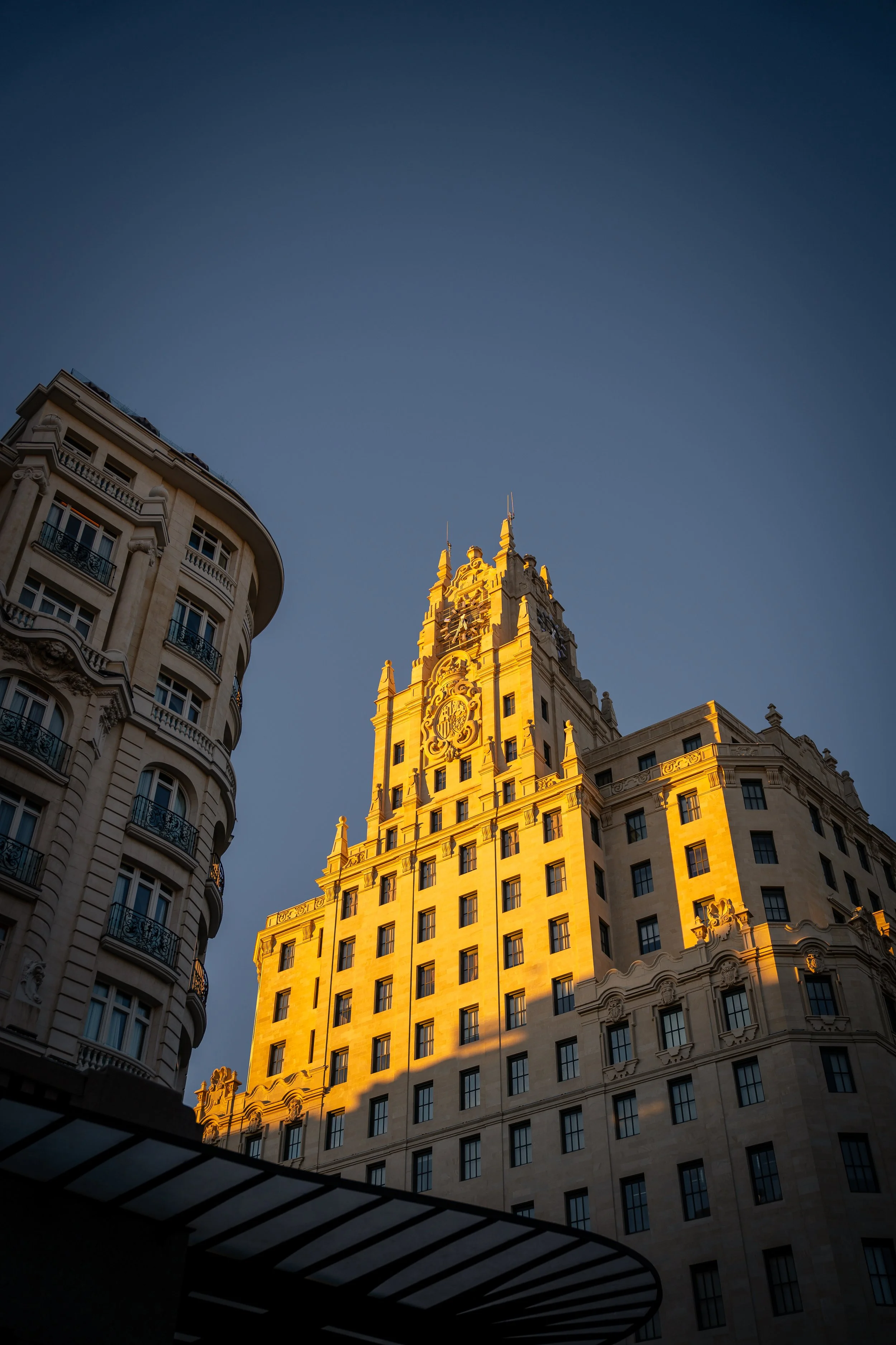 Ein heller, historischer Wolkenkratzer mit gotischer Architektur im Sonnenlicht, umgeben von anderen Gebäuden, unter einem klaren blauen Himmel.