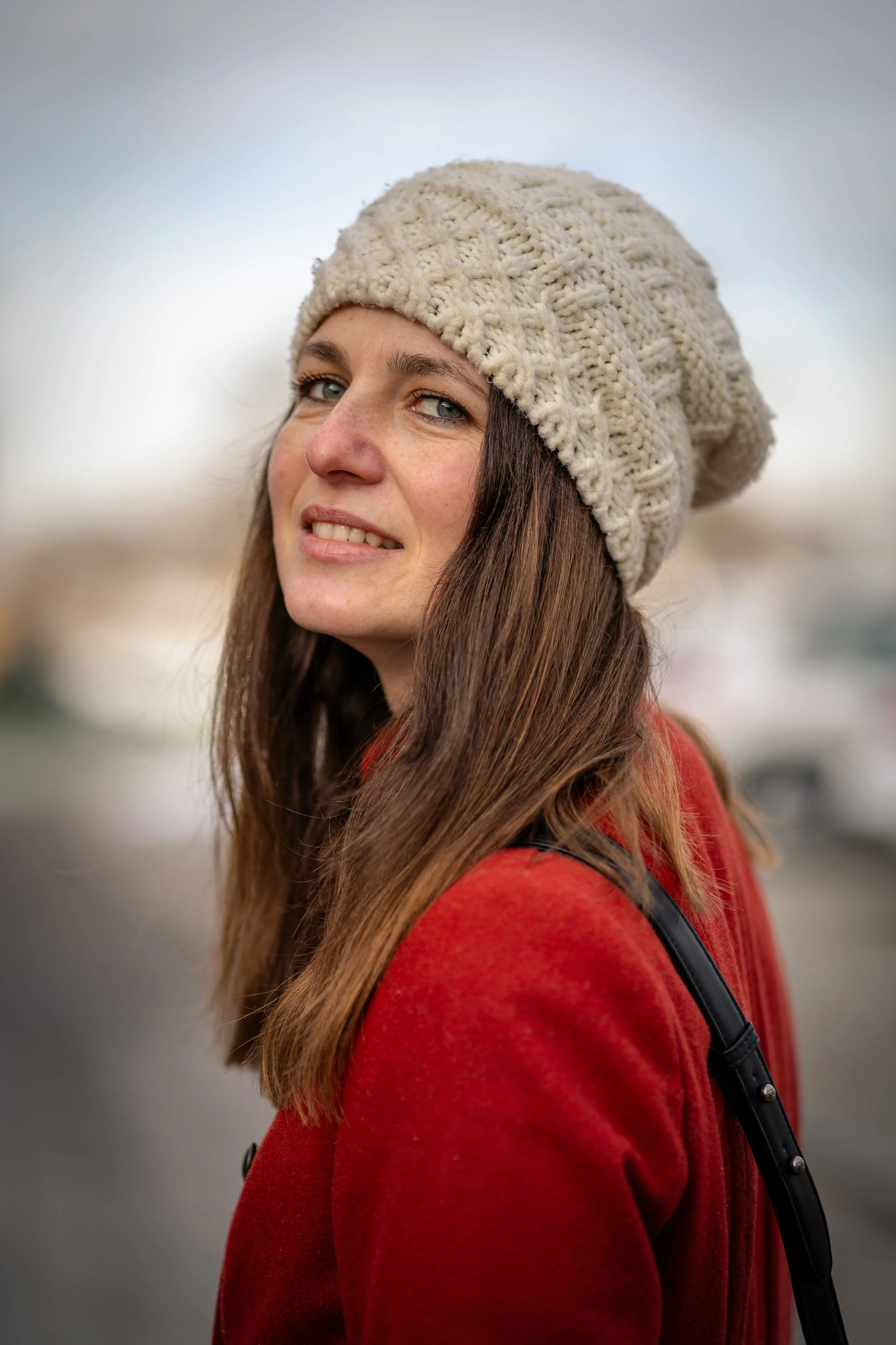 Junge Frau mit braunen Haaren, trägt eine graffiti-weiße Wollmütze und roten Mantel, steht vor einem grau-blauen Himmel, lächelt leicht, trägt einen schwarzen Rucksack.