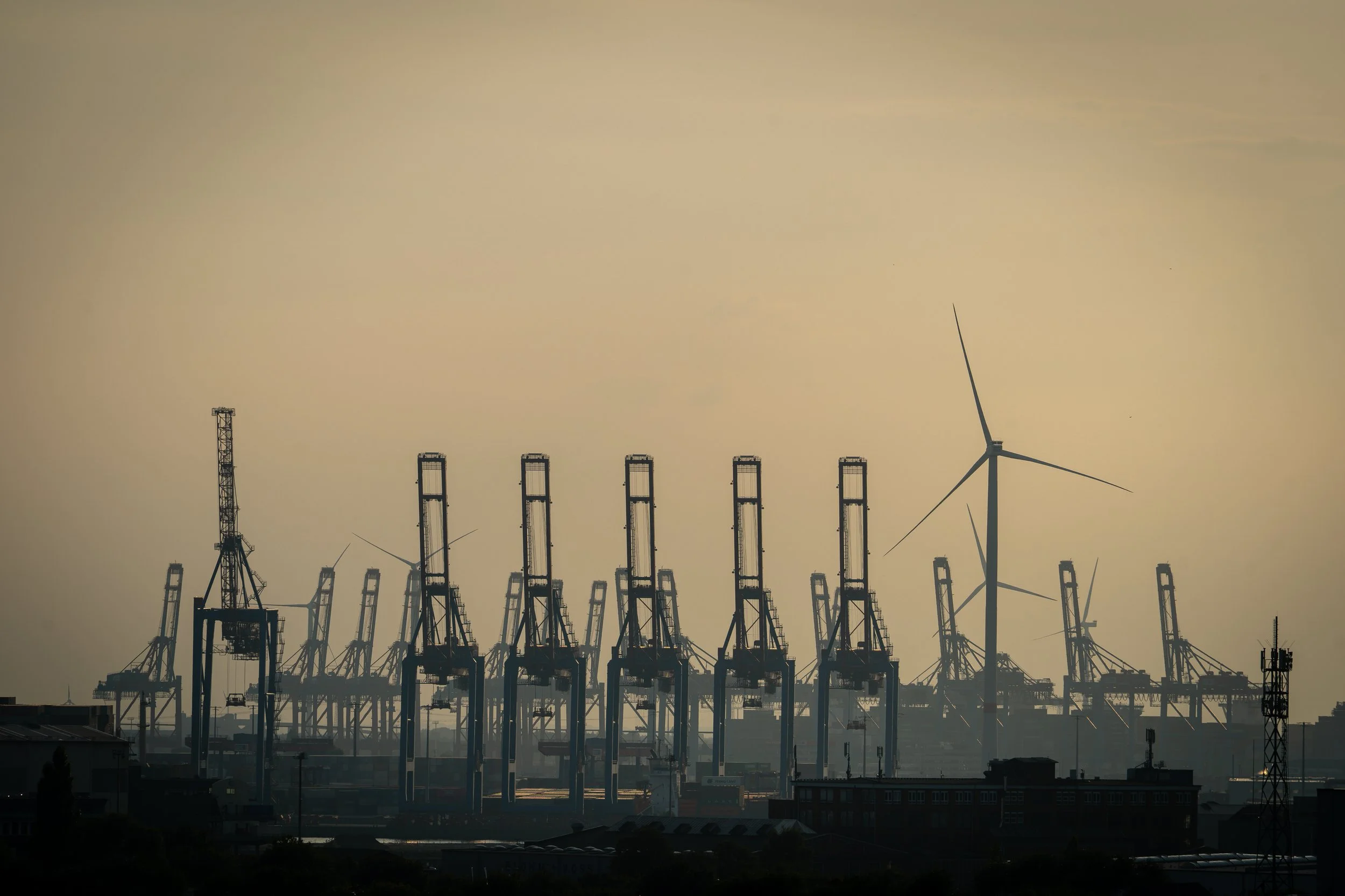 Blick auf eine Offshore-Windkraftanlage und Containerkräne bei Sonnenuntergang, im Vordergrund dunkle Gebäude