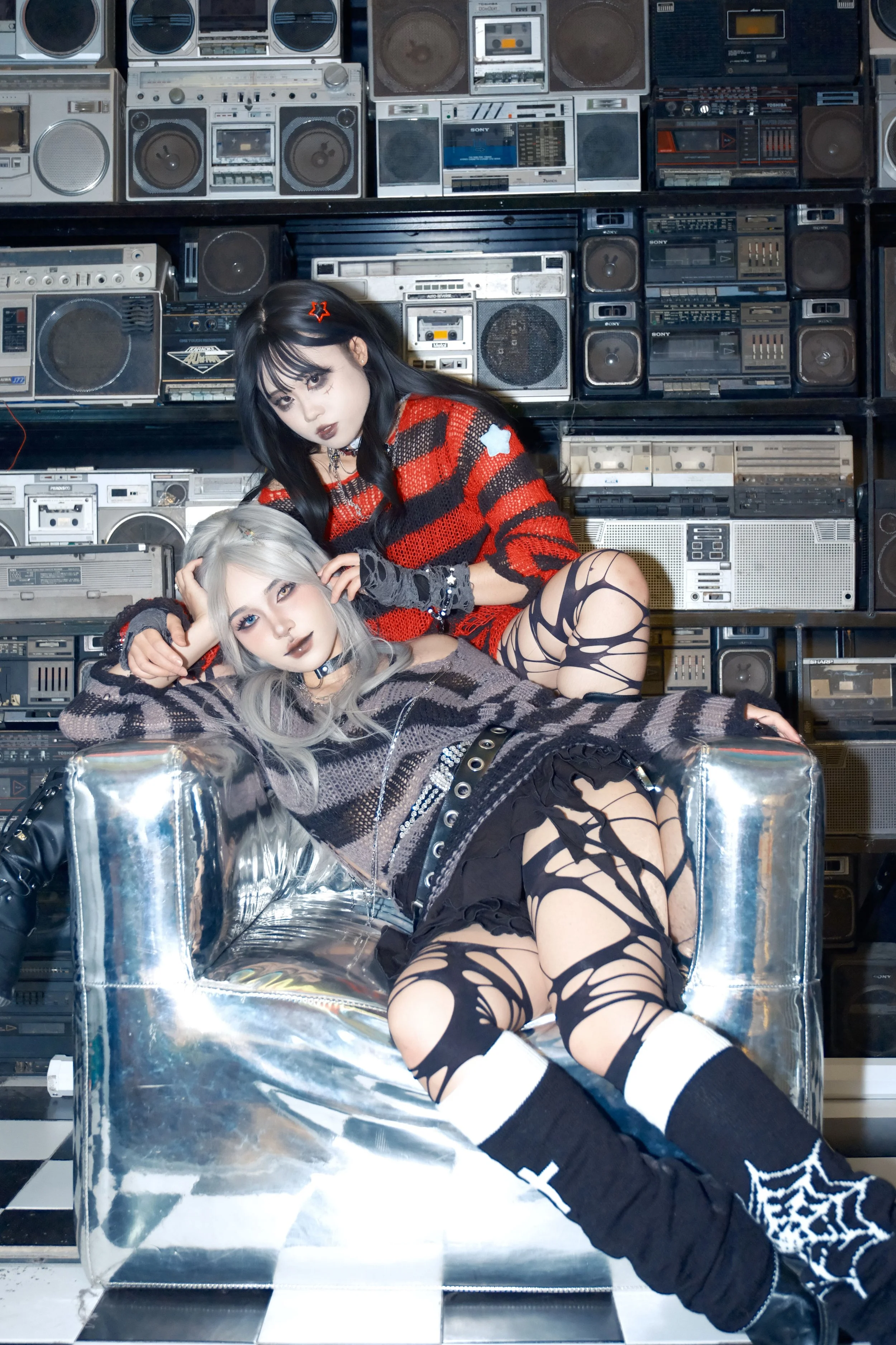 Two women sitting on a bright silver couch, wearing stripey knitted jumpers and ripped tights with vintage stereos in the background