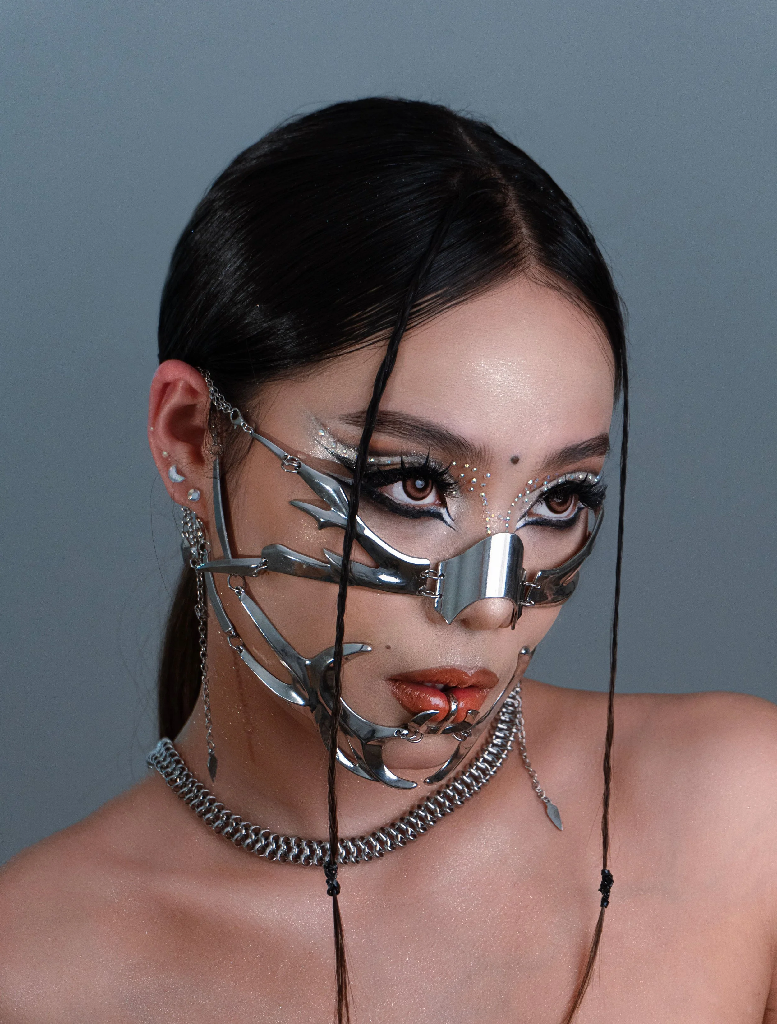 Headshot of a woman with two face framing braids and silver gemstones and graphic eye makeup. She is wearing a chunky silver chain necklace and cyber nose plate and lip piercing.