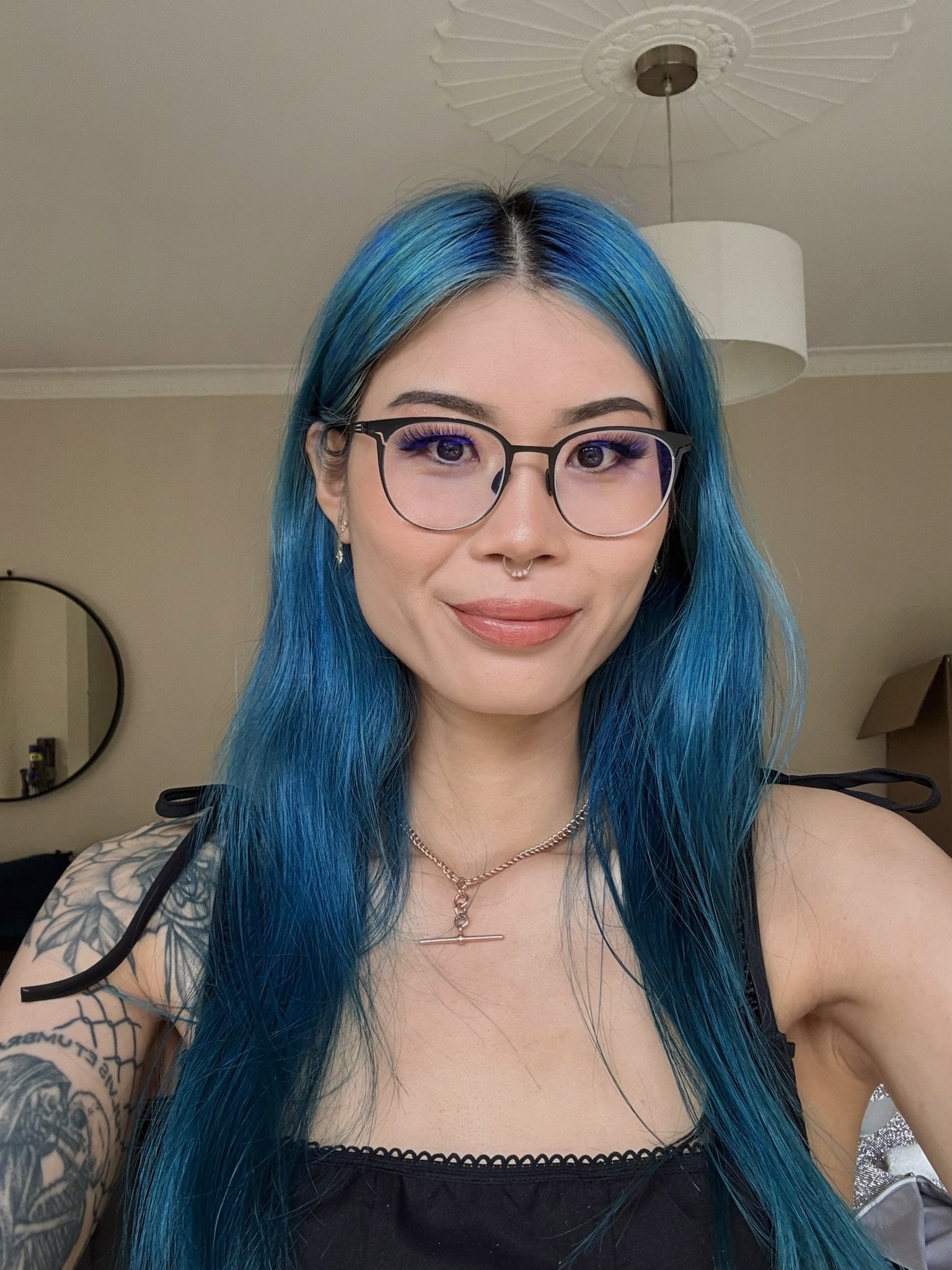Young East Asian woman with long blue hair and glasses, taking a selfie in natural lighting and smiling into the camera