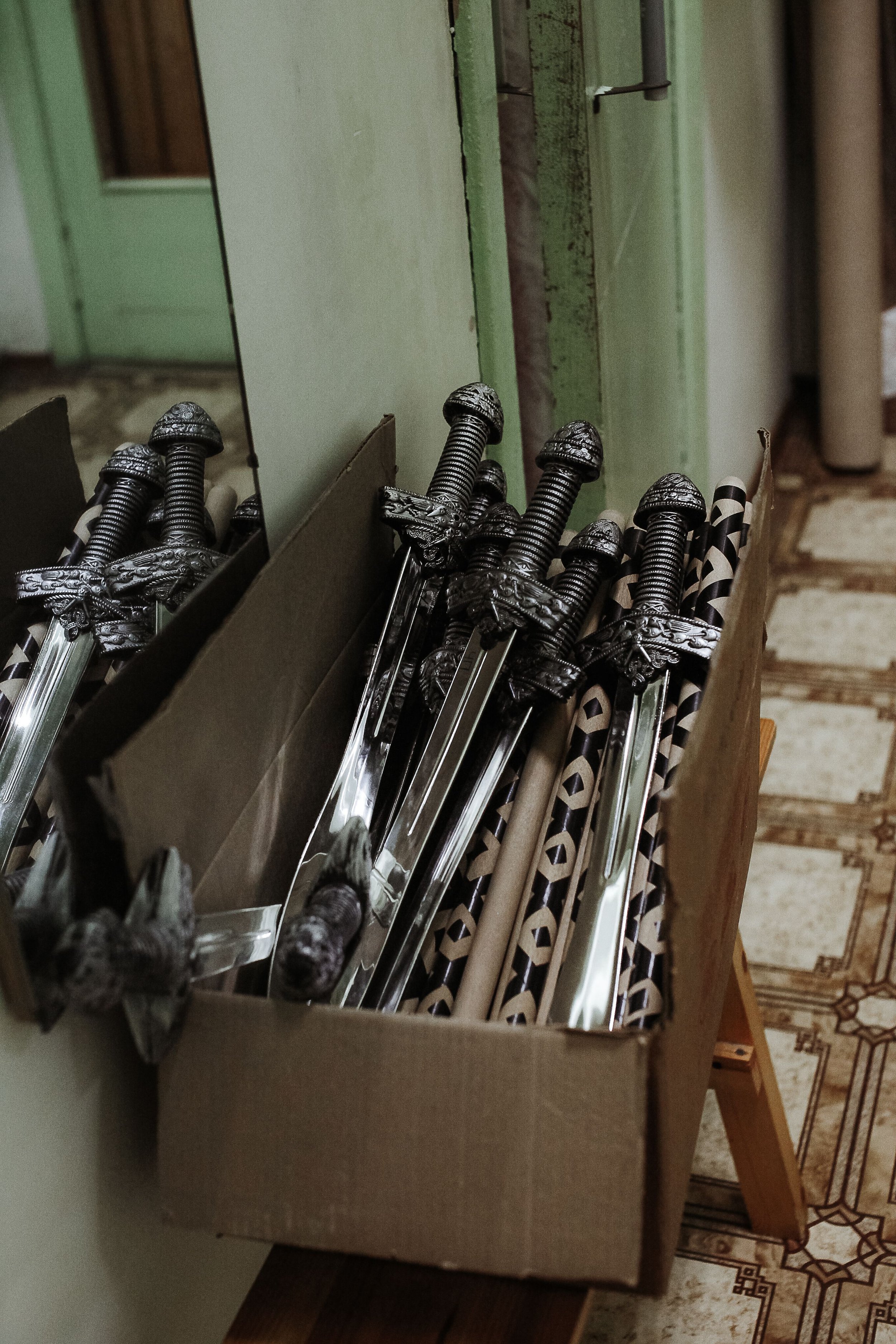 A cardboard box filled with decorative swords, some with black and metallic handles, placed on a patterned tiled floor next to a wooden support and a wall with a door in the background.