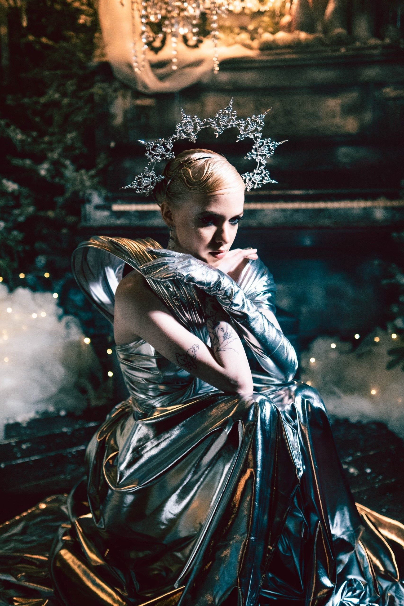 A woman with platinum blonde hair styled in finger waves, wearing a shiny silver metallic dress and long gloves, sitting and resting her chin on her hand in a festive, dimly lit setting with fairy string lights.
