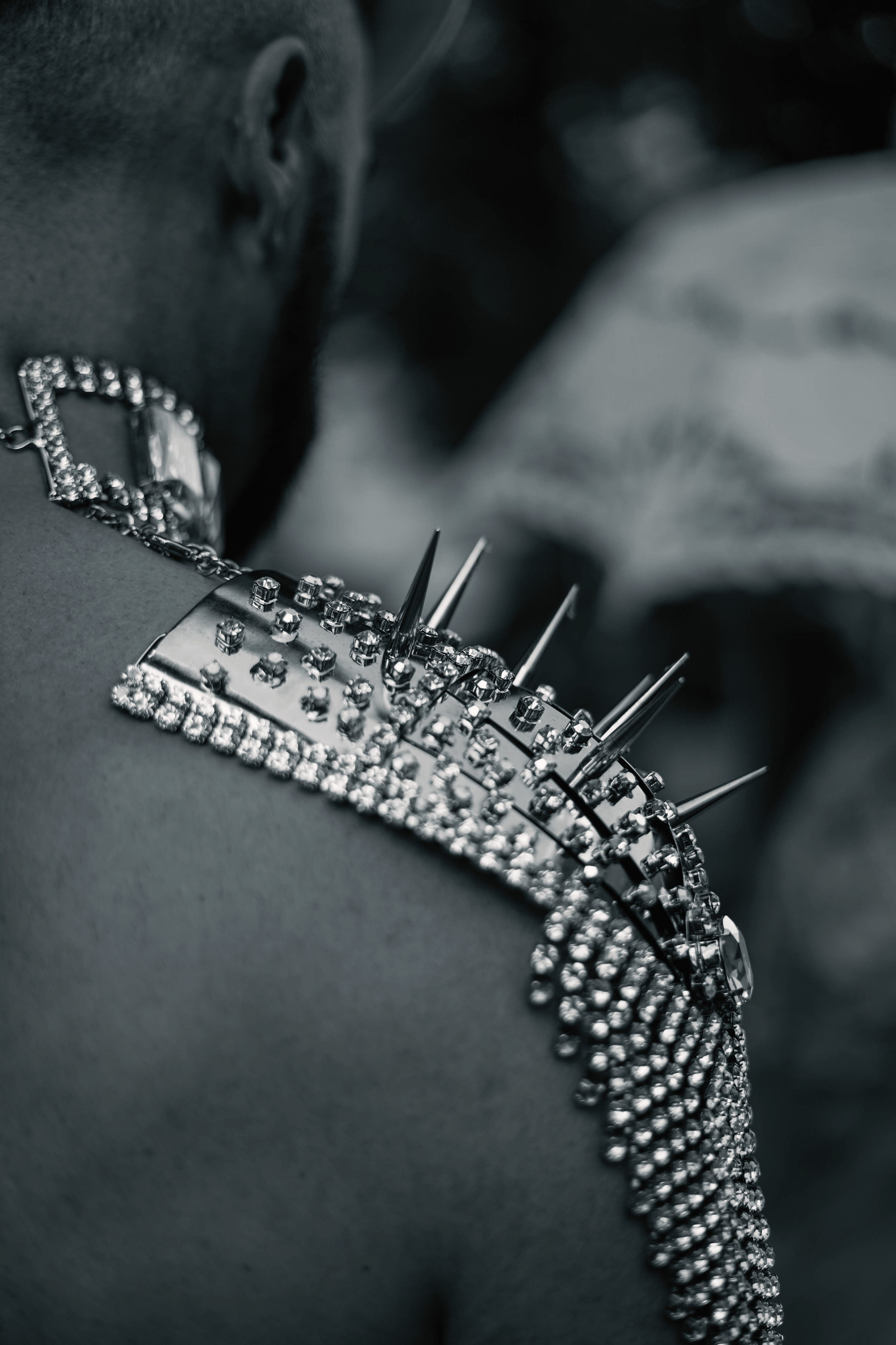Black and white photo of a shirtless man's right shoulder, wearing an ornamental metal epaulet spaulder with diamantes and decorative spikes and studs