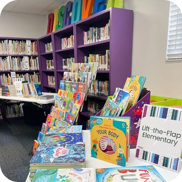 Book display with colorful children's books in front of purple bookshelves in an elementary school library.