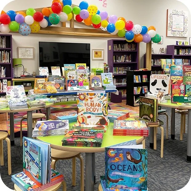Colorful balloons decorate the ceiling of a children's bookstore or library with books displayed on round tables and shelves, and a large TV mounted on the wall.