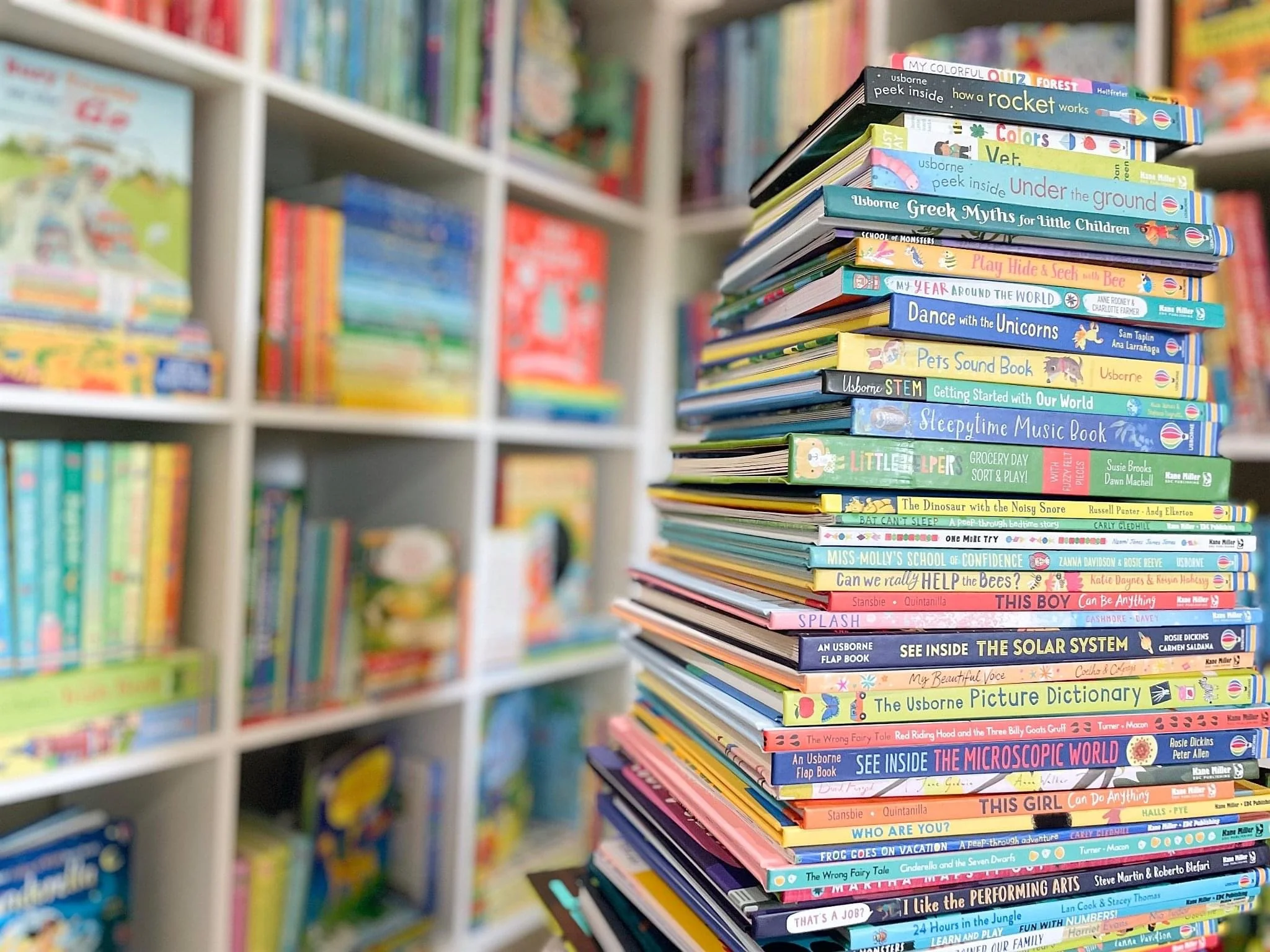 A close-up of a colorful stack of children's books on a table in a bookstore, with shelves of more books blurred in the background.