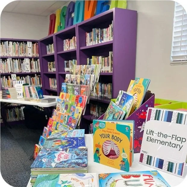 Bookshelf in a library with children's books on display, colorful signs reading 'Science', and a sign indicating it's at Lift-the-Flap Elementary.