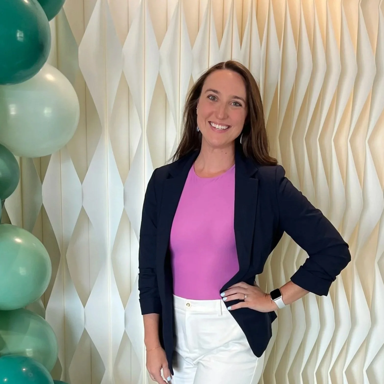 A woman with long brown hair smiling and standing in front of a cream-colored geometric 3D wall with large teal and white balloons to the left. She is wearing a dark blazer over a pink top, white pants, a smartwatch, and earrings.