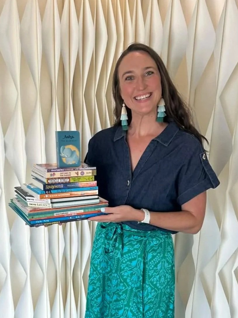 A woman with long brown hair wearing teal earrings, a dark blue shirt, and teal patterned pants, smiling and holding a stack of books in front of a cream-colored textured curtain.