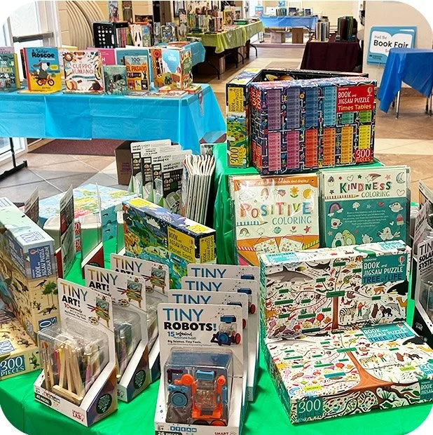 A display of books, puzzles, coloring books, and educational toys at a book fair or sale event, with tables covered in blue and green cloths.