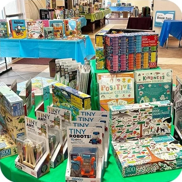 Display of children's books, puzzles, coloring books, and toys at a book fair, with tables covered in colorful tablecloths.
