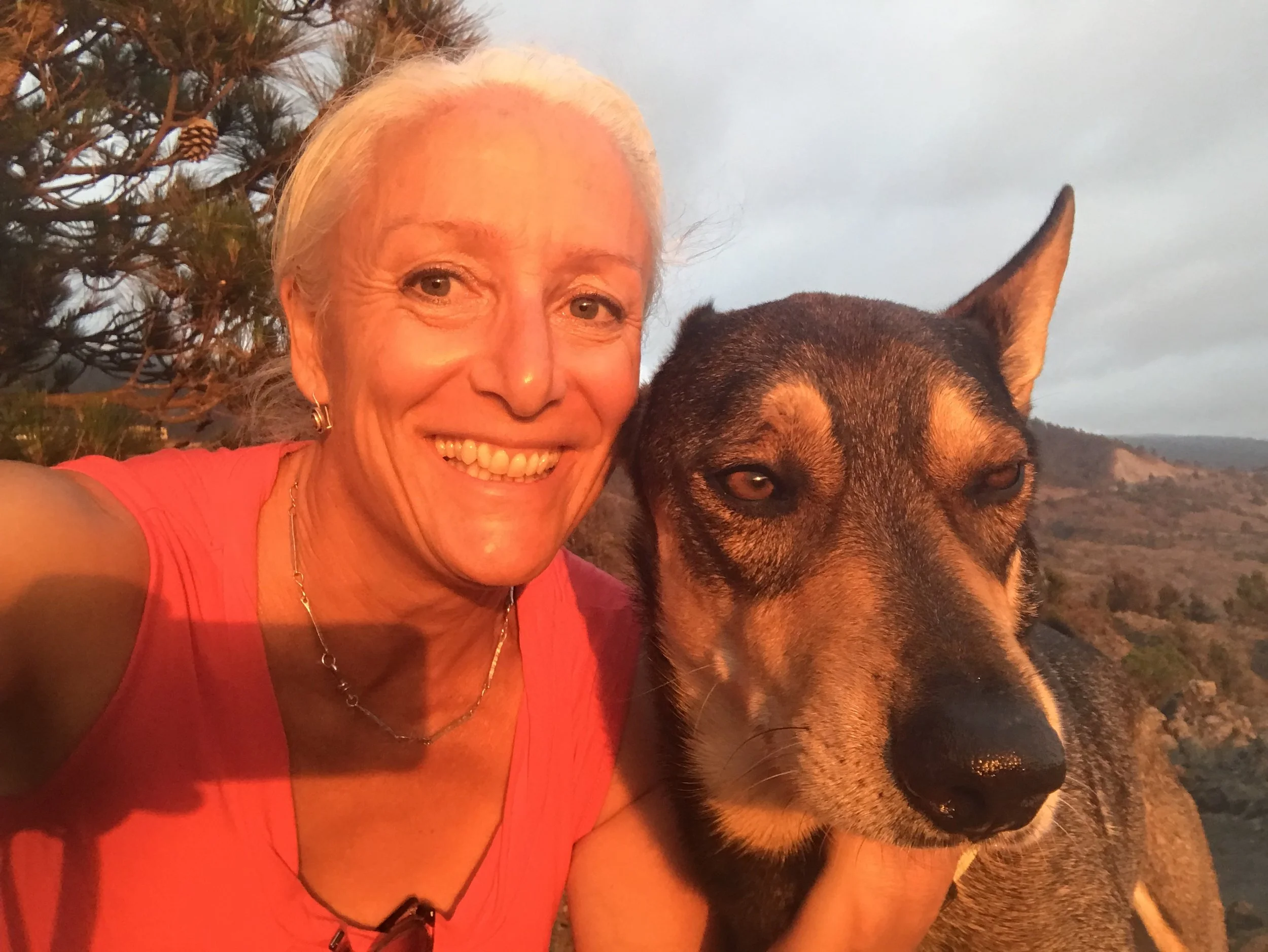 Mujer mayor sonriendo con un perro pastor alemán en un paisaje al atardecer en un entorno natural con árboles y colinas.