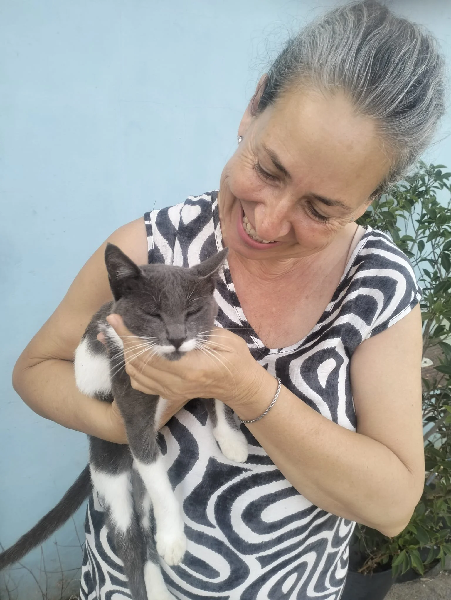 Mujer con cabello canoso sosteniendo un gato gris y blanco, sonriendo en un fondo de pared azul y plantas.