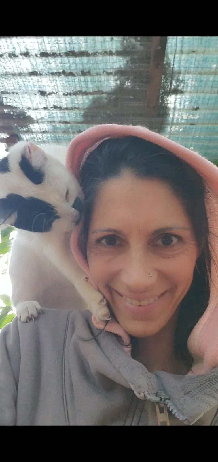 Una mujer con sonrisa, lleva una capucha rosada y una chaqueta gris, con un gato negro y blanco sobre su hombro izquierdo, en un entorno al aire libre con sombra y estructura metálica en el fondo.
